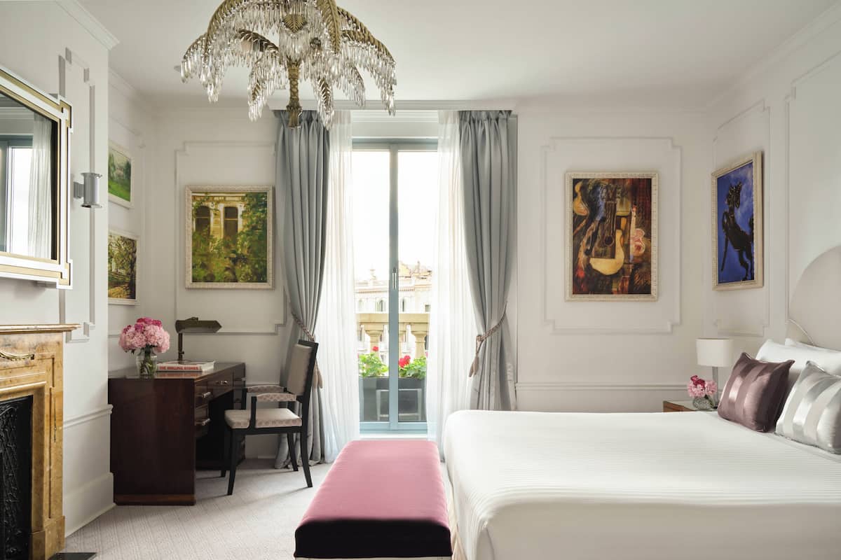 A hotel room with a bed, a desk, a chair, a fireplace, a chandelier, and artwork on the walls.