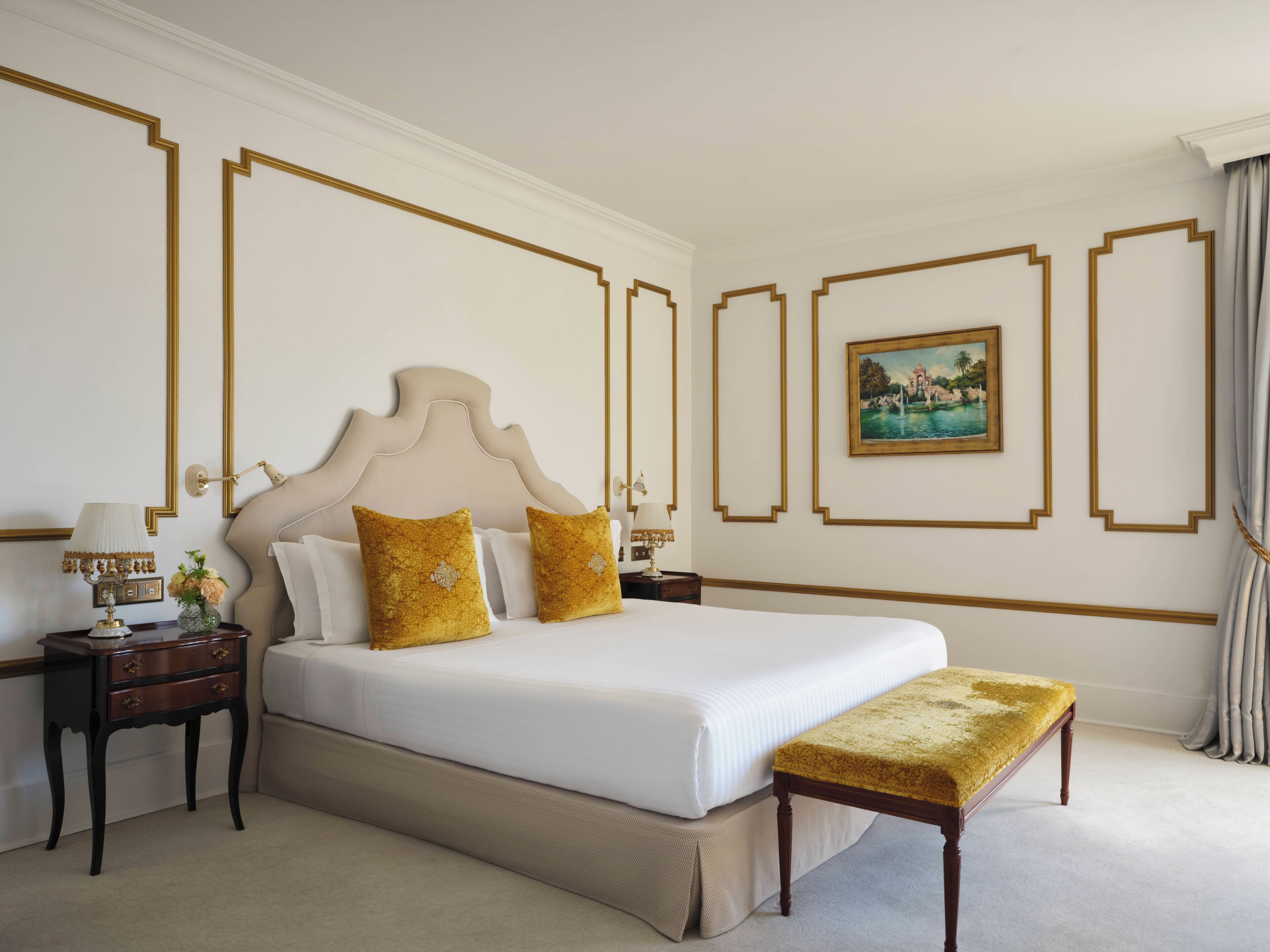 A hotel room with a large bed, a bedside table with a lamp, a bench, and a framed picture on the wall.