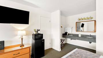 A hotel room with a wooden dresser, a black refrigerator, a flat-screen TV, a vanity with a mirror, and a bed with a patterned bedspread.