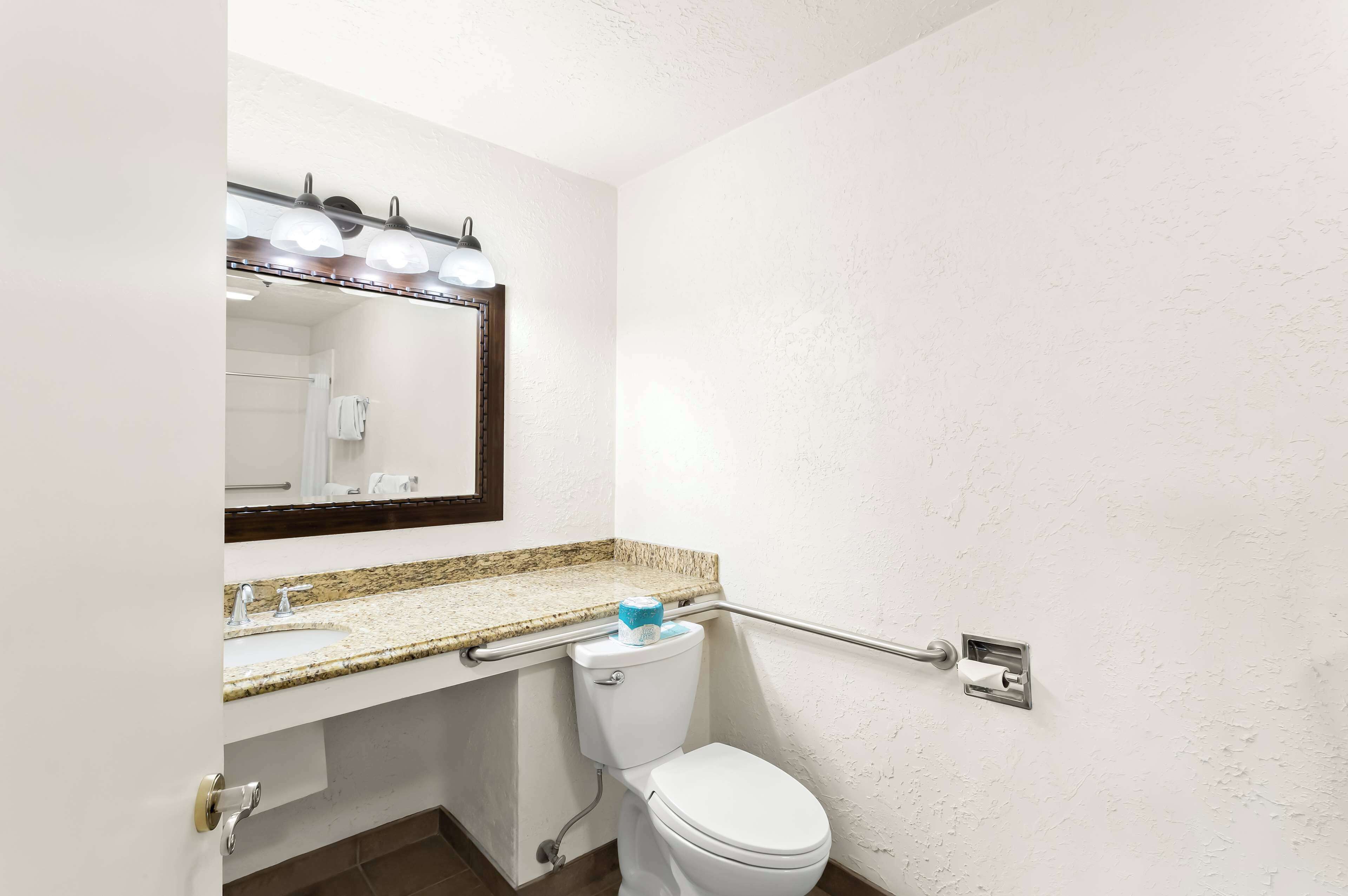 A bathroom with a white toilet, a granite countertop sink, a mirror with lights, and a grab bar.