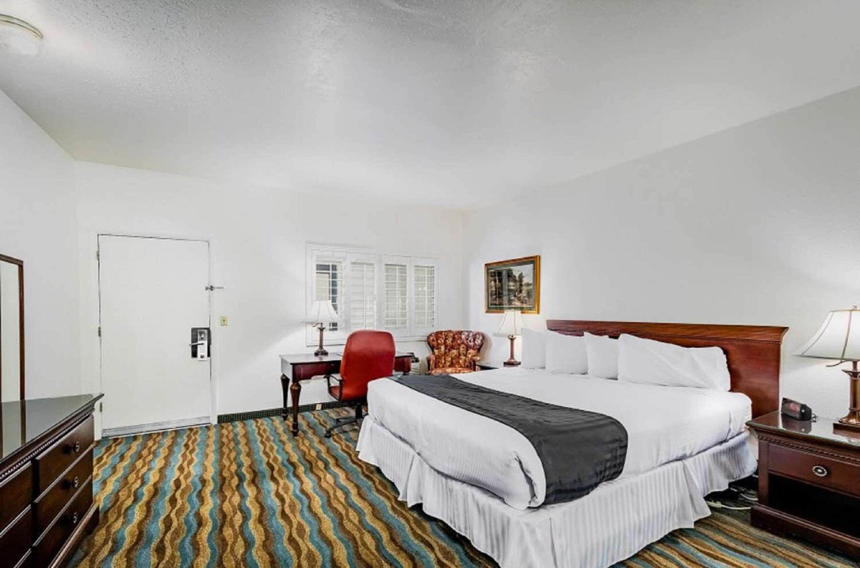 Un cuarto de hotel con una cama grande, un escritorio con silla, un sillón rojo, una lámpara de escritorio y un cuadro en la pared.
