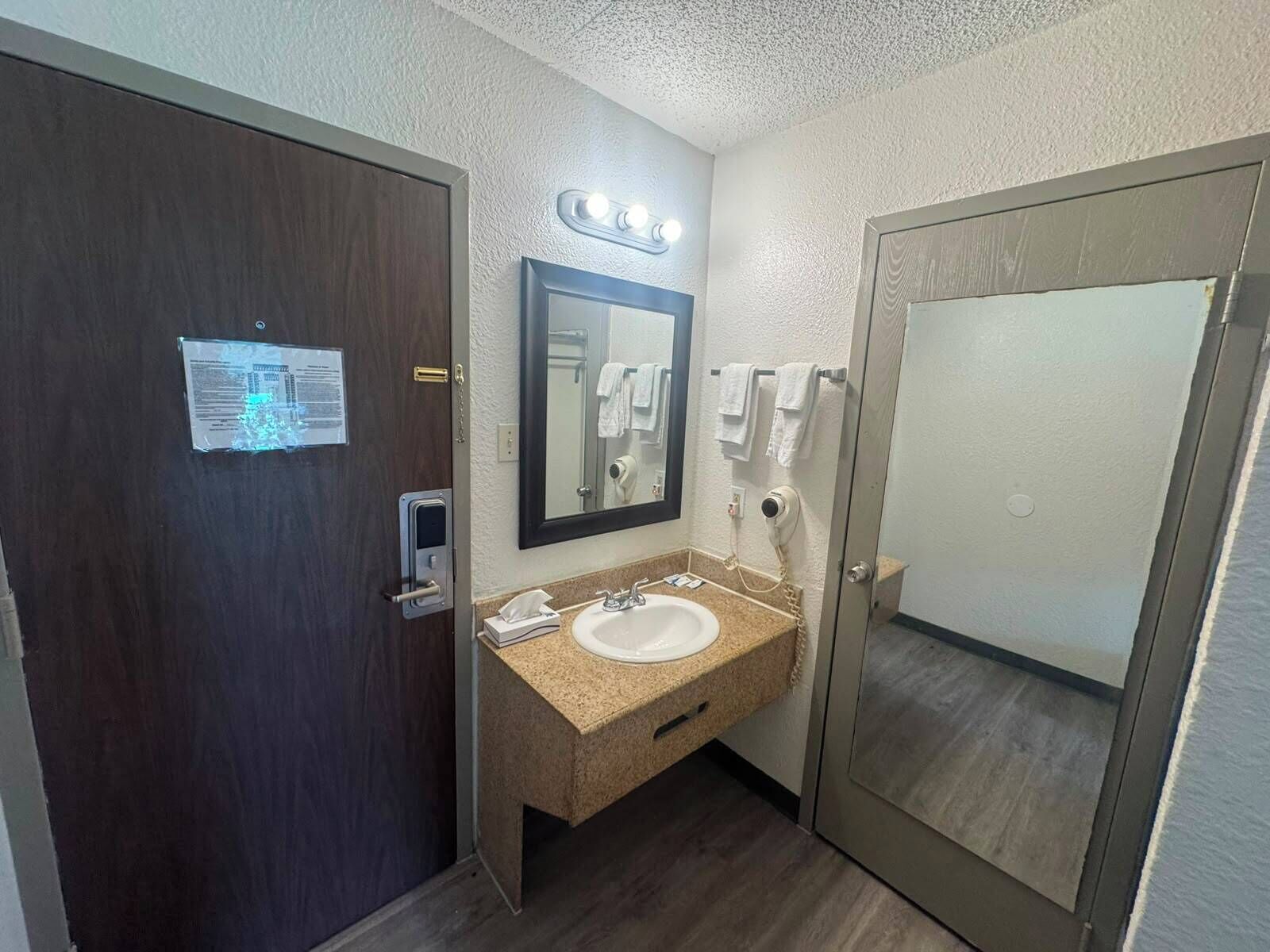 A hotel bathroom with a sink, mirror, towel rack, and a door with a sign.