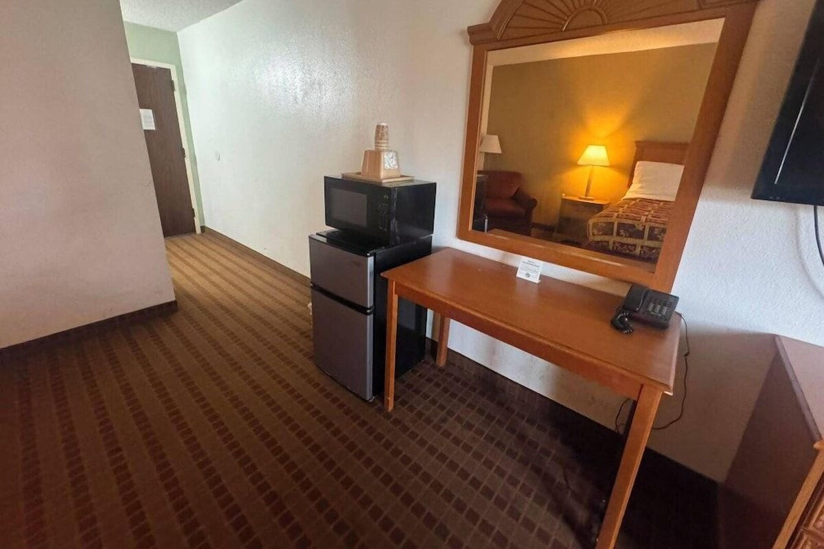 A hotel room with a desk, a television, a refrigerator, a mirror, and a lamp.