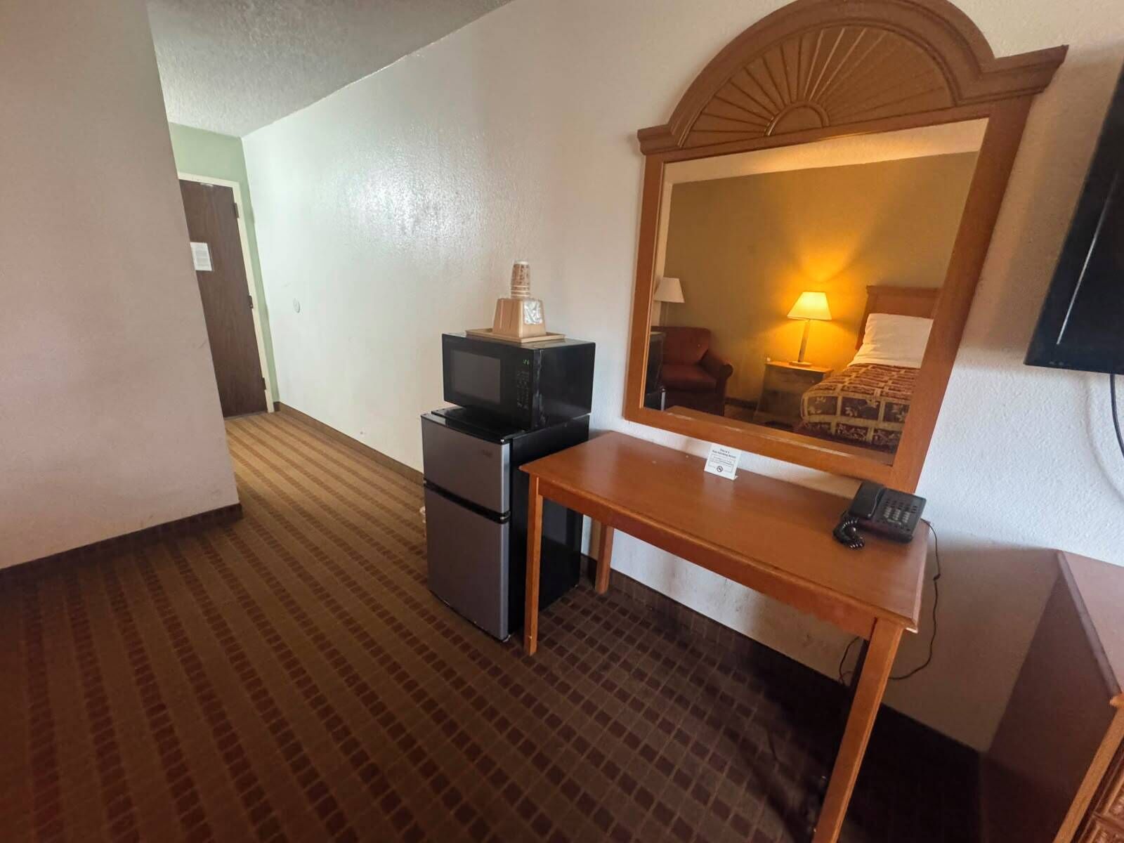 A hotel room with a kitchenette, a wooden desk, a telephone, and a large mirror.