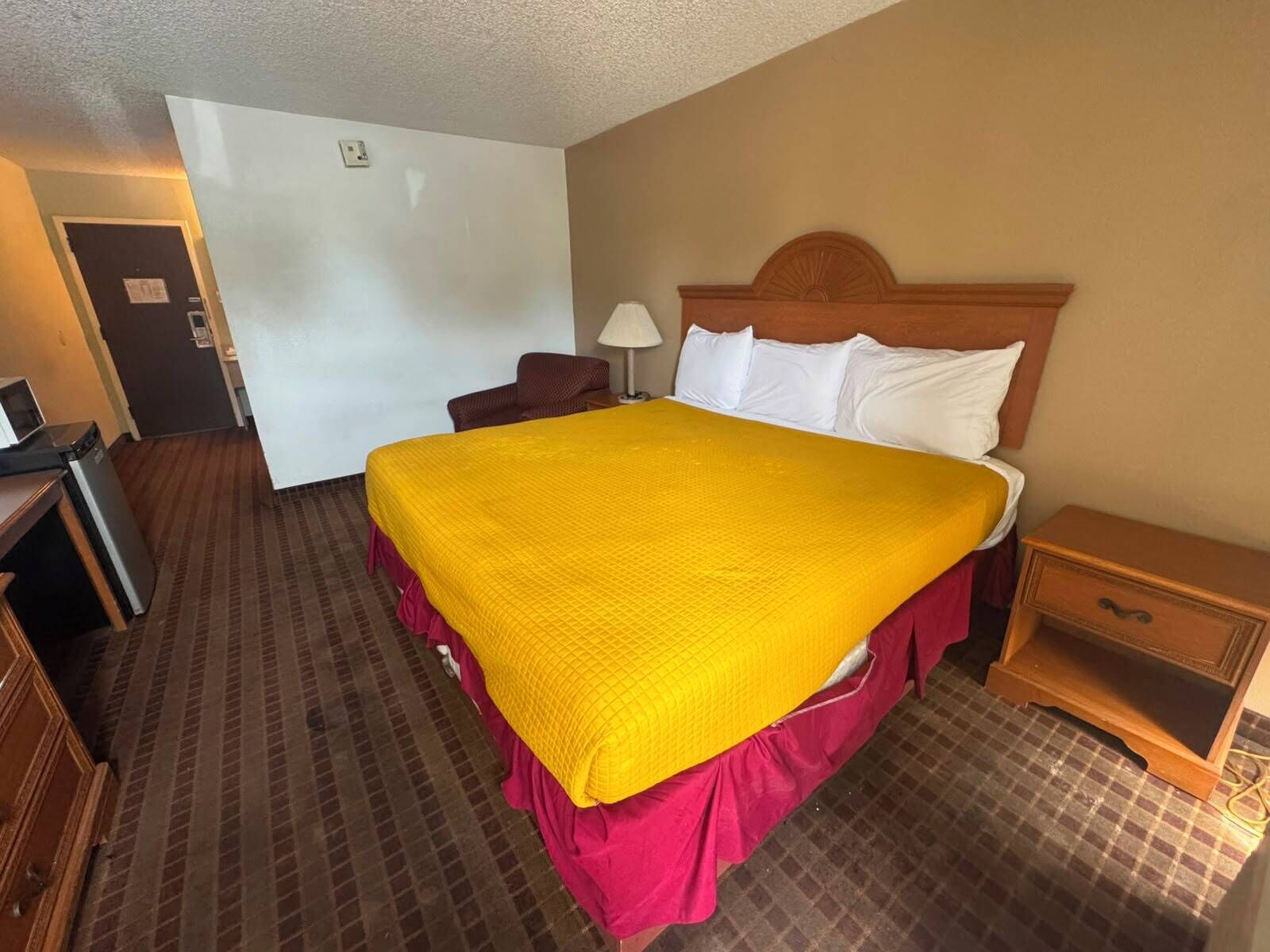 A hotel room with a large bed, a wooden headboard, a desk with a lamp, and a small wooden nightstand.