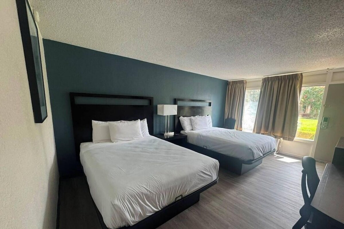 A hotel room with two beds, a desk, and a view of the outdoors.