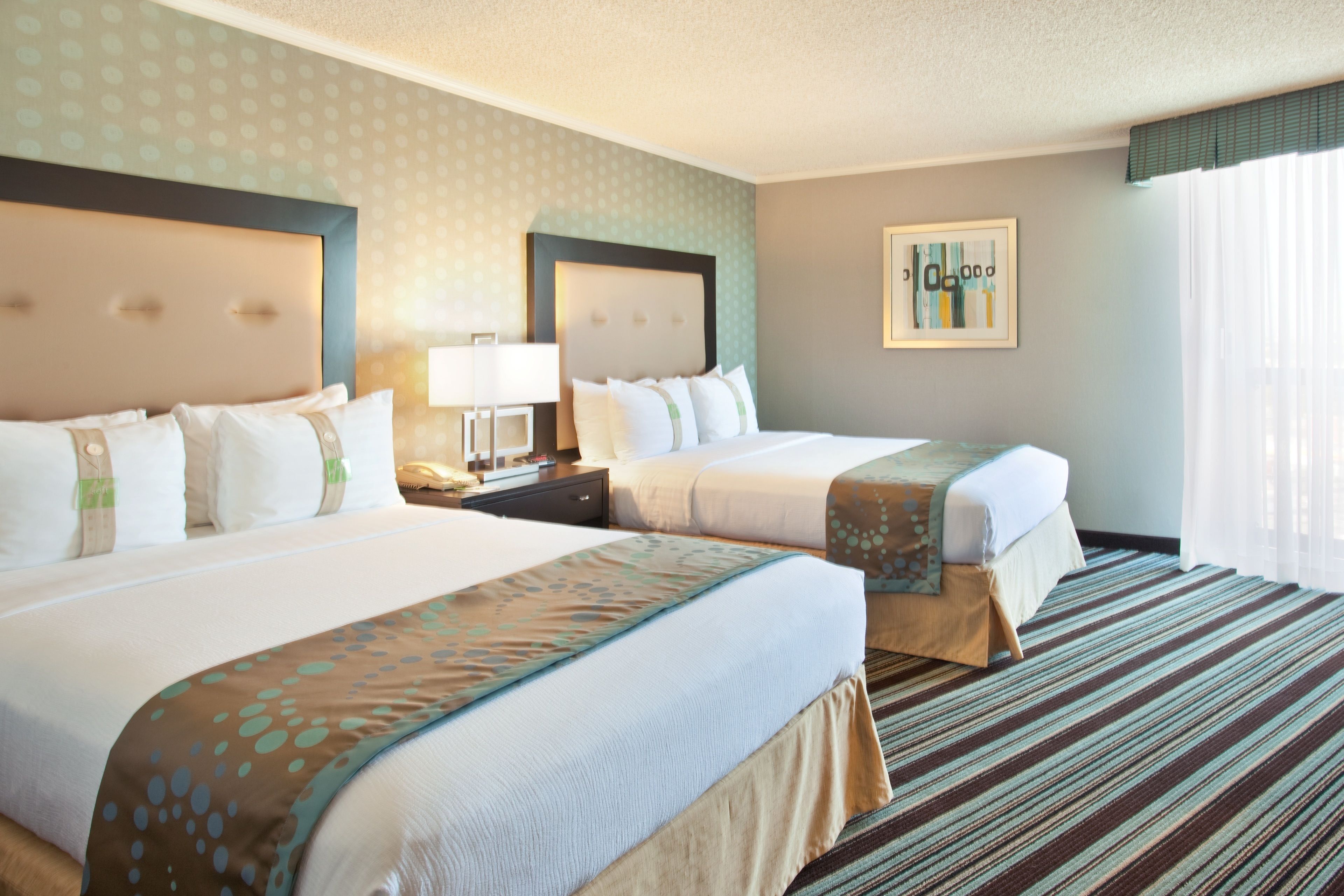 A hotel room with two beds, a nightstand, a lamp, a framed picture on the wall, and a window with curtains.