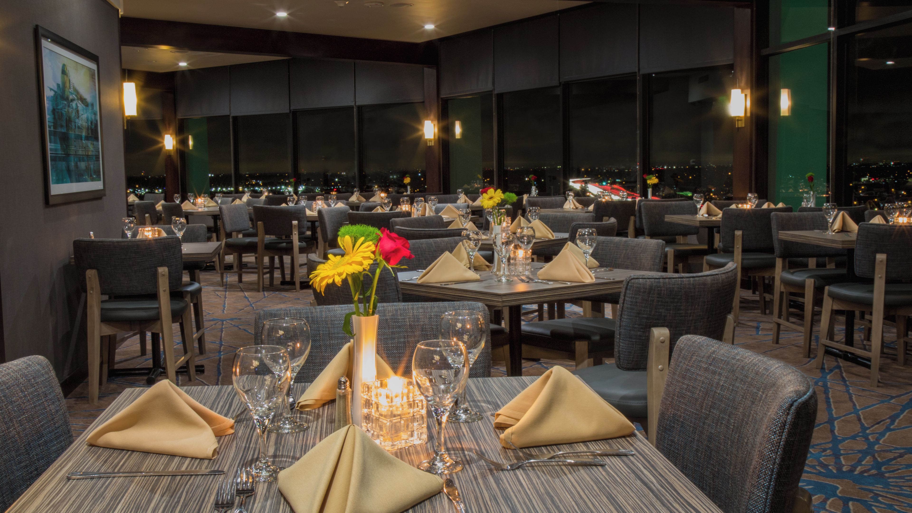 Un restaurant doté de nombreuses tables, de chaises et offrant une vue sur la ville grâce à de grandes fenêtres.