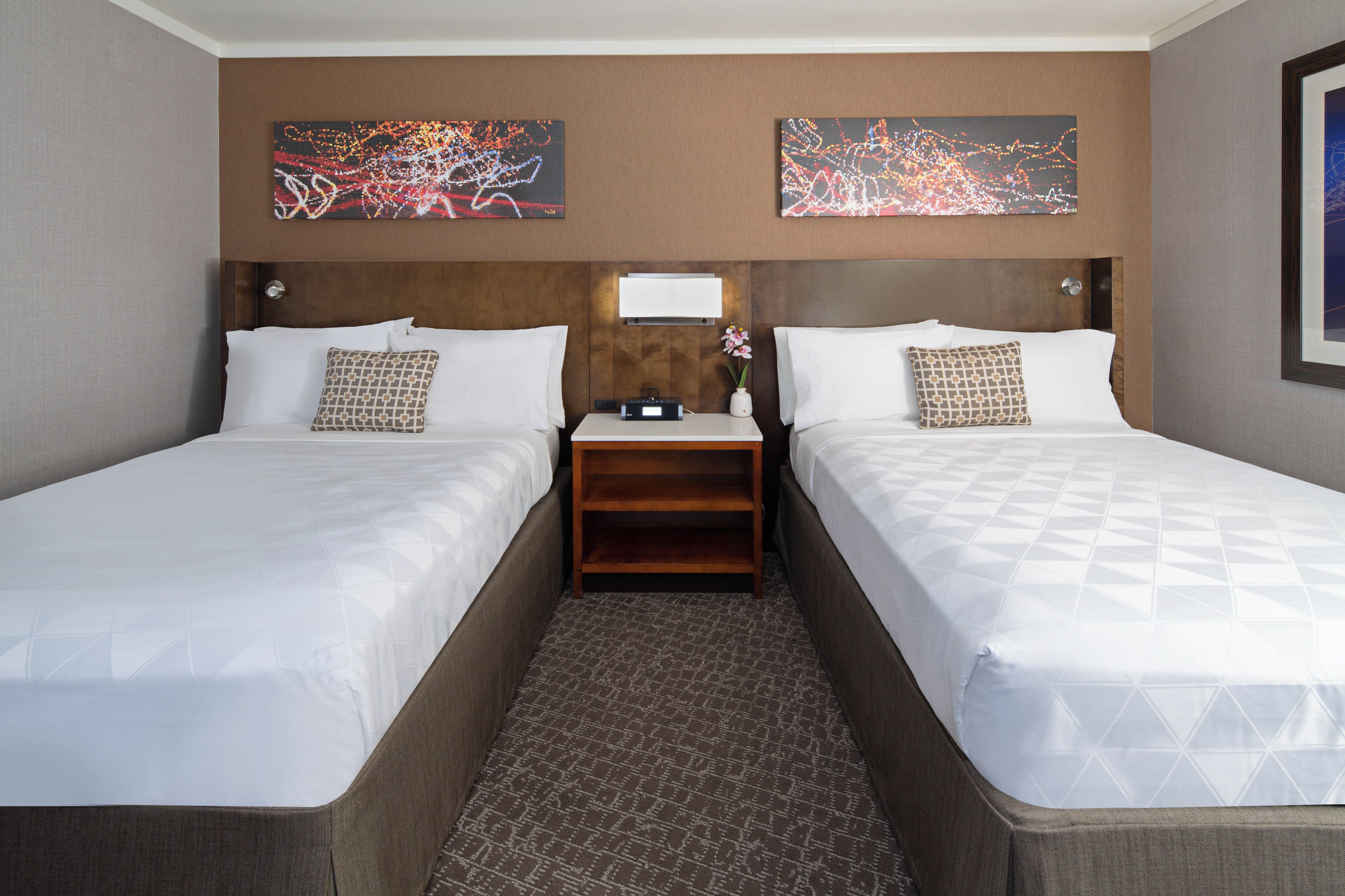 A hotel room with two beds, a nightstand, a mirror, and two abstract paintings on the wall.