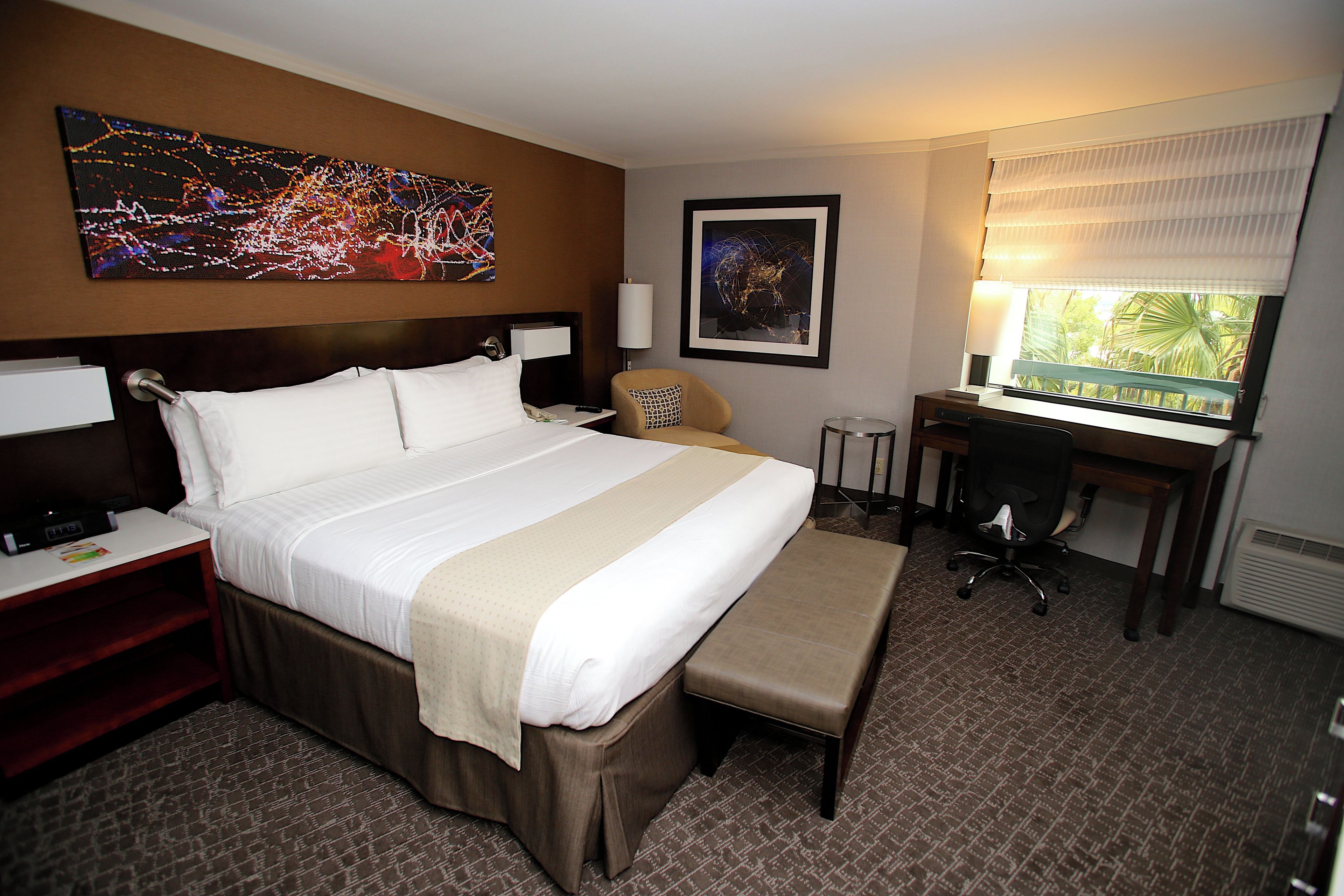 A hotel room with a large bed, a desk, a chair, a bench, a TV, a lamp, and artwork on the walls.