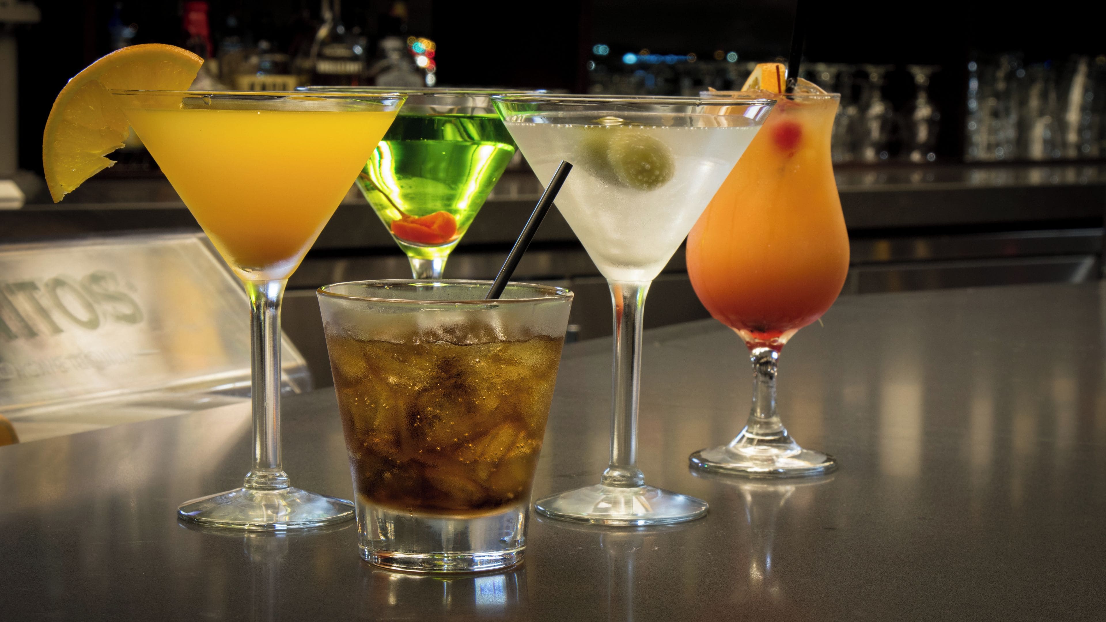 A bar with various drinks, including cocktails and a soda, with a bar counter and bottles in the background.