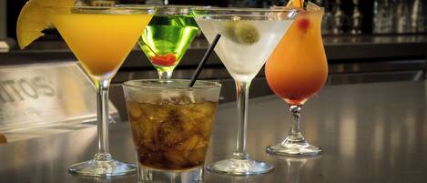 A bar with various drinks, including cocktails and a soda, with a bar counter and bottles in the background.
