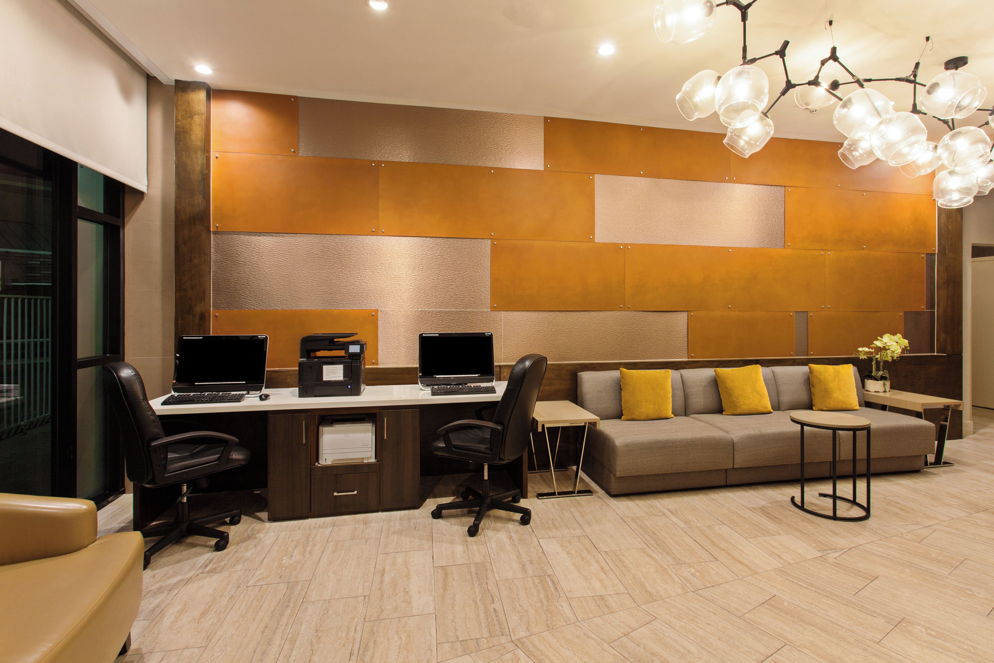 Un espace de bureau moderne comprenant un bureau, deux ordinateurs portables, une imprimante, un canapé avec des coussins jaunes et un luminaire au plafond au design original.