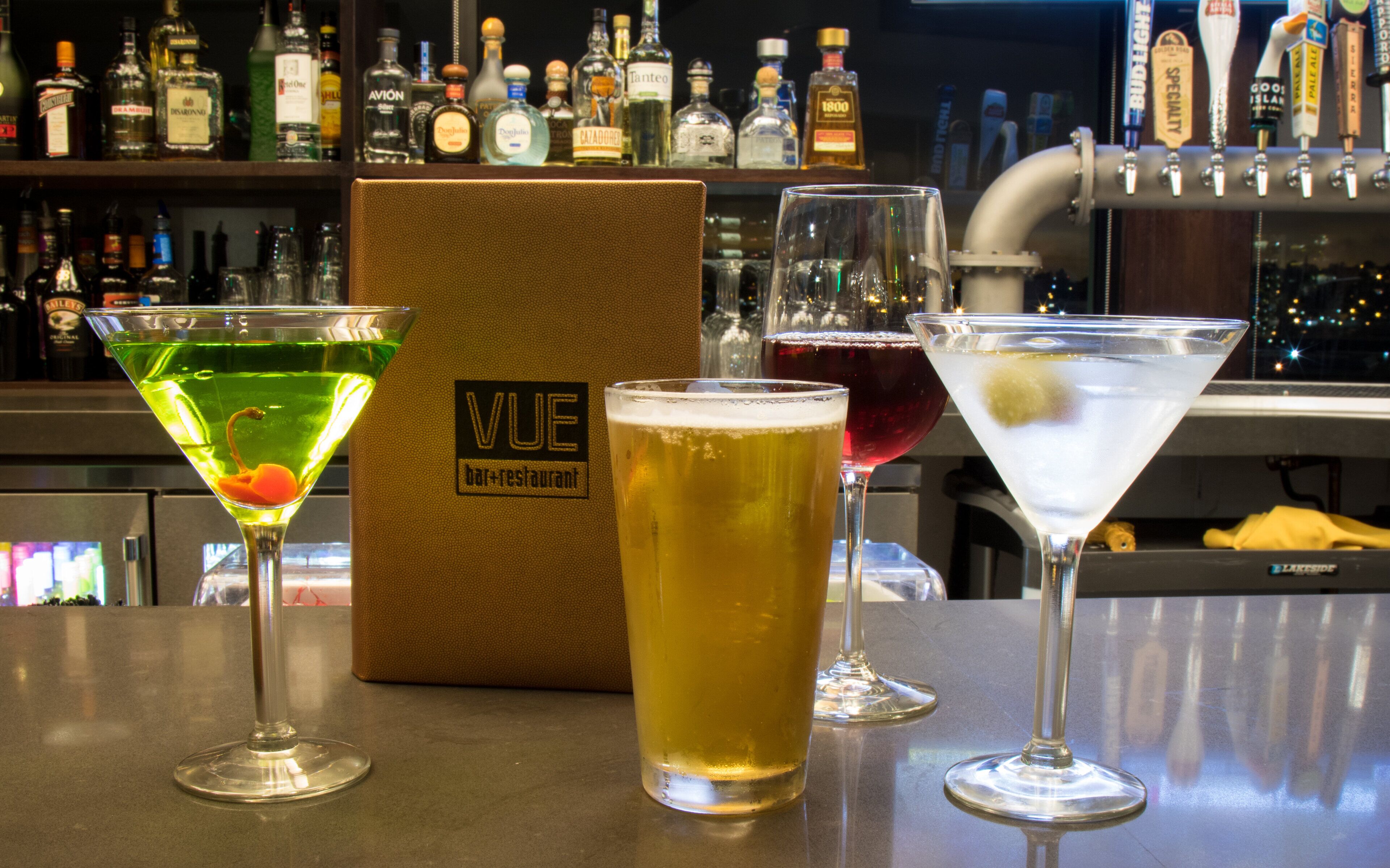 Three drinks on a bar counter with a bottle of VUE bar+restaurant in the background.