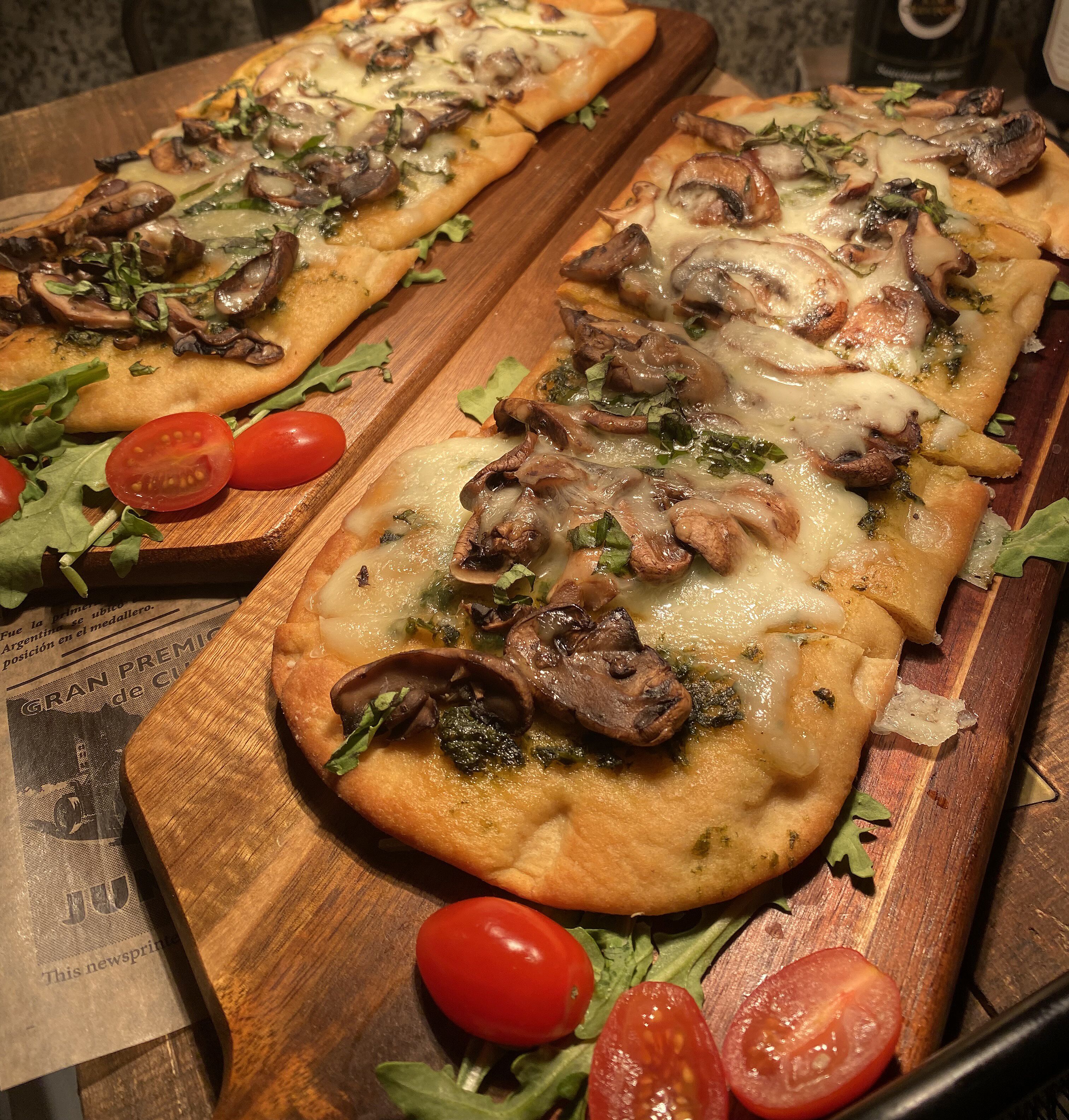 Dos pizzas sobre tablas de madera, con tomates y verduras de hoja verde.