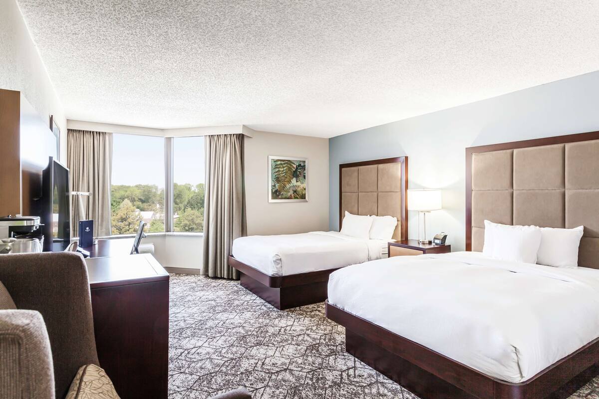 A hotel room with two beds, a desk, a TV, and a large window with curtains.