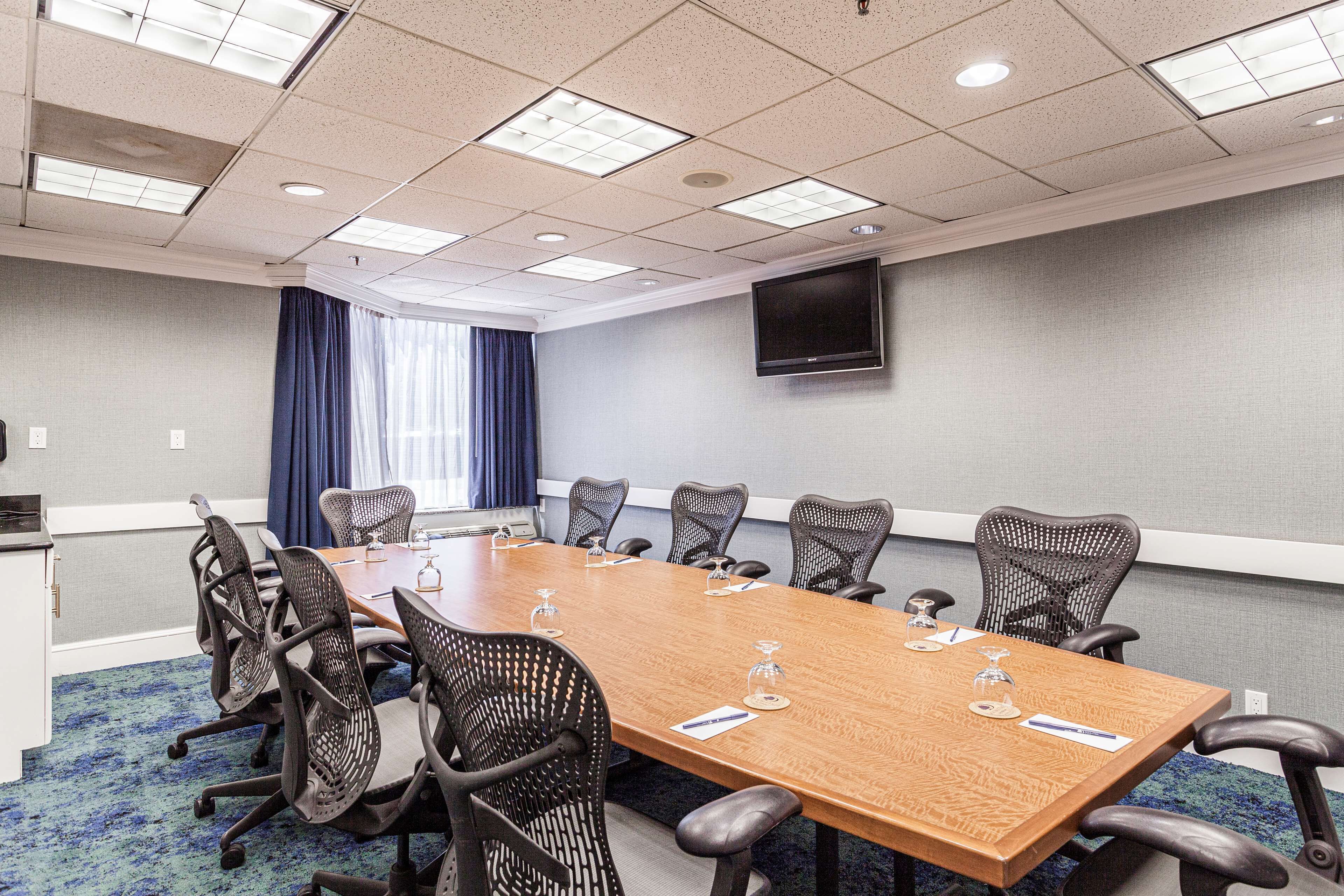 A conference room with a long table, chairs, a flat-screen TV, and a window with curtains.