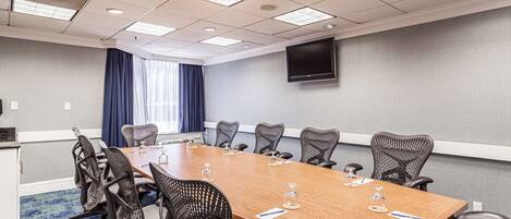 A conference room with a long table, chairs, a flat-screen TV, and a window with curtains.