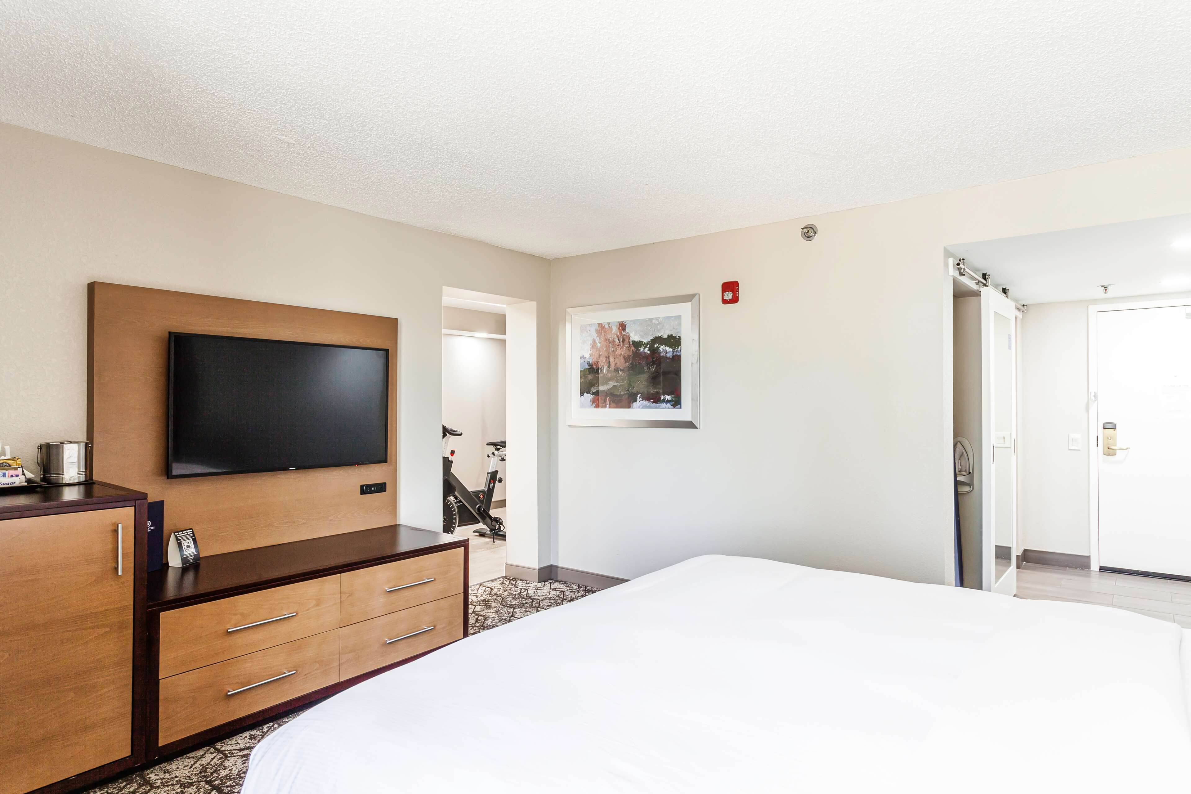 A hotel room with a large bed, a flat-screen TV mounted on a wooden cabinet, a framed picture on the wall, and a door leading to another room.