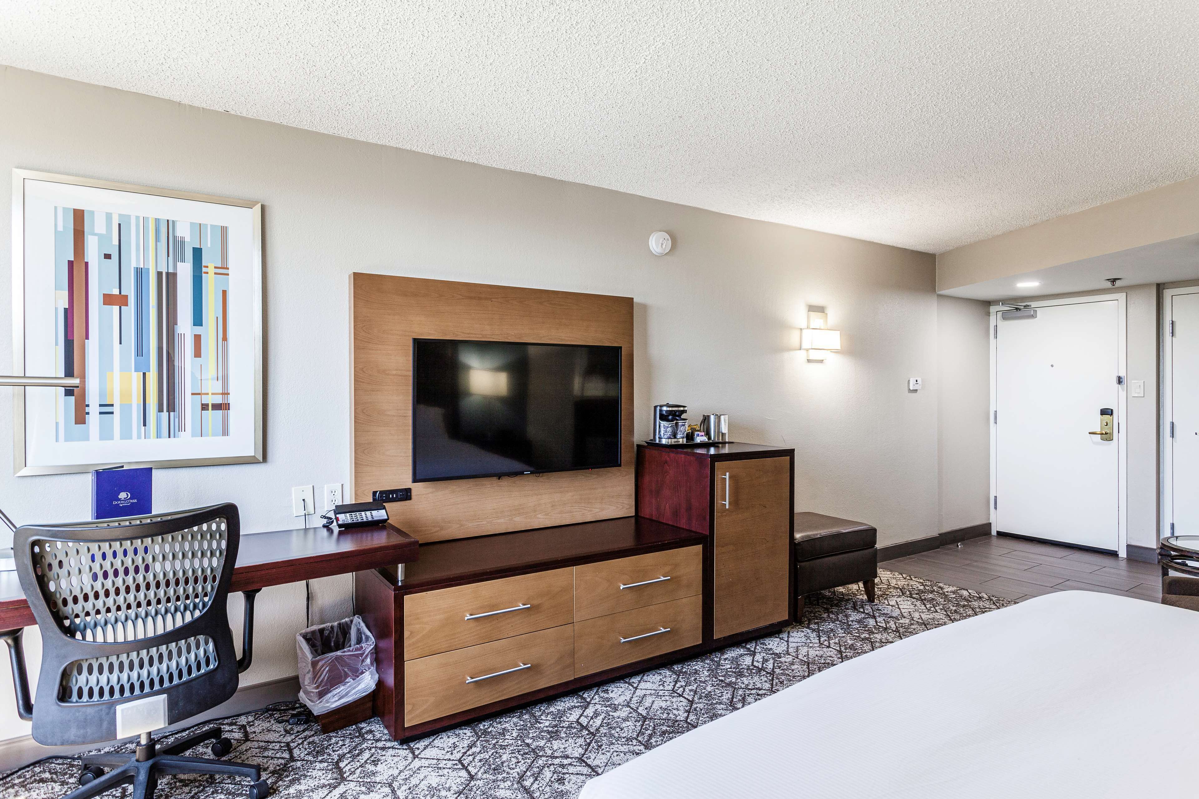 A hotel room with a bed, a desk with a computer, a chair, a TV mounted on the wall, and a cabinet.