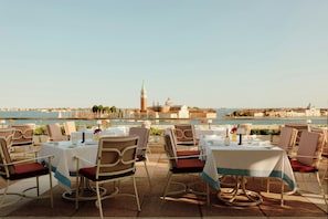An outdoor dining area with tables and chairs, overlooking a body of water and historic buildings.