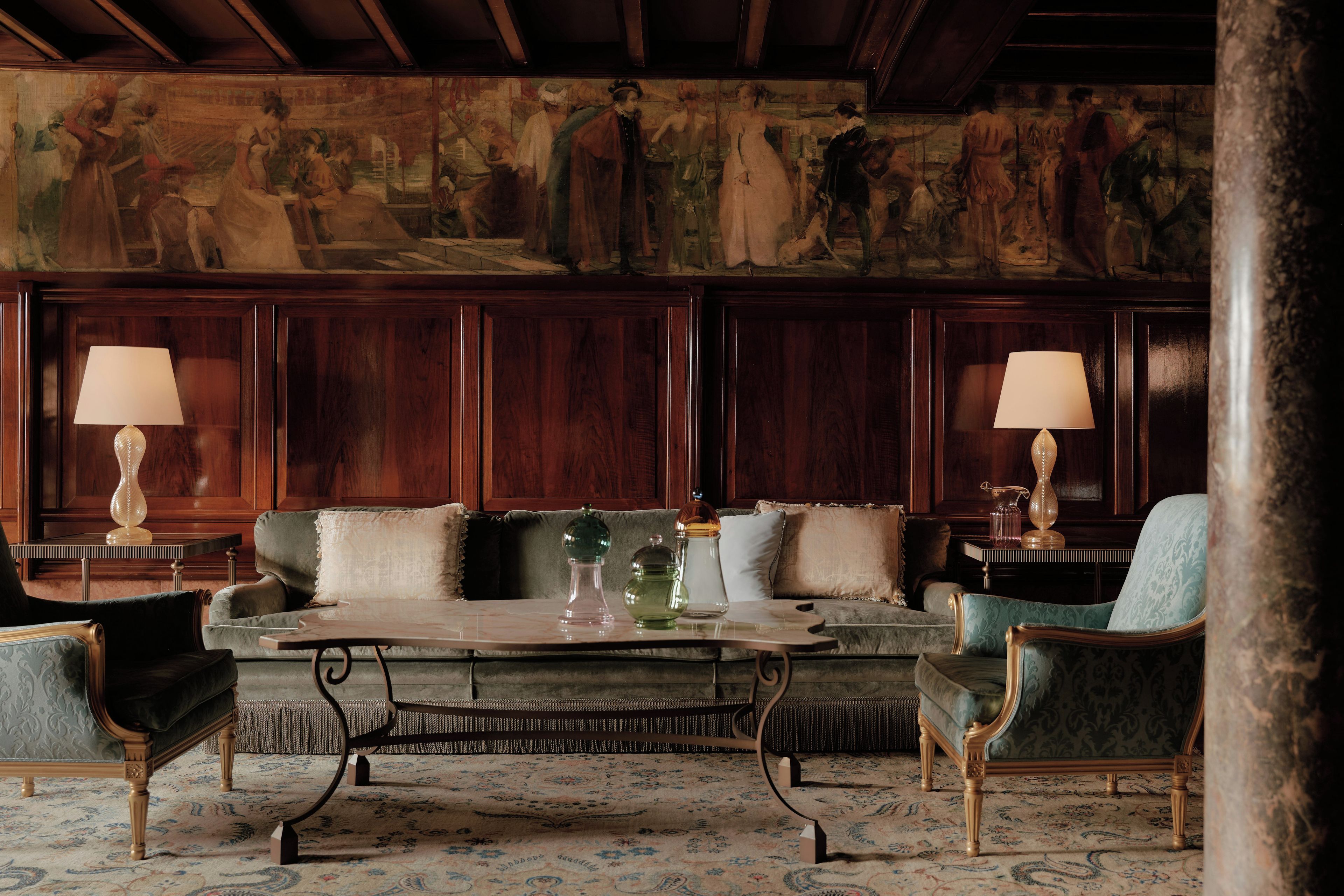 A room with a sofa, two armchairs, a coffee table, and lamps. A large painting is on the wall.