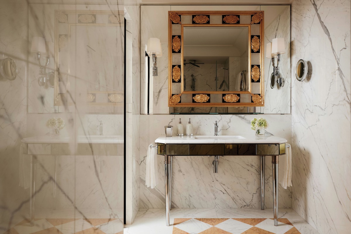 Un bagno con pavimento in marmo, uno specchio grande con cornice decorativa, un lavabo con rubinetteria cromata e due luci a parete.