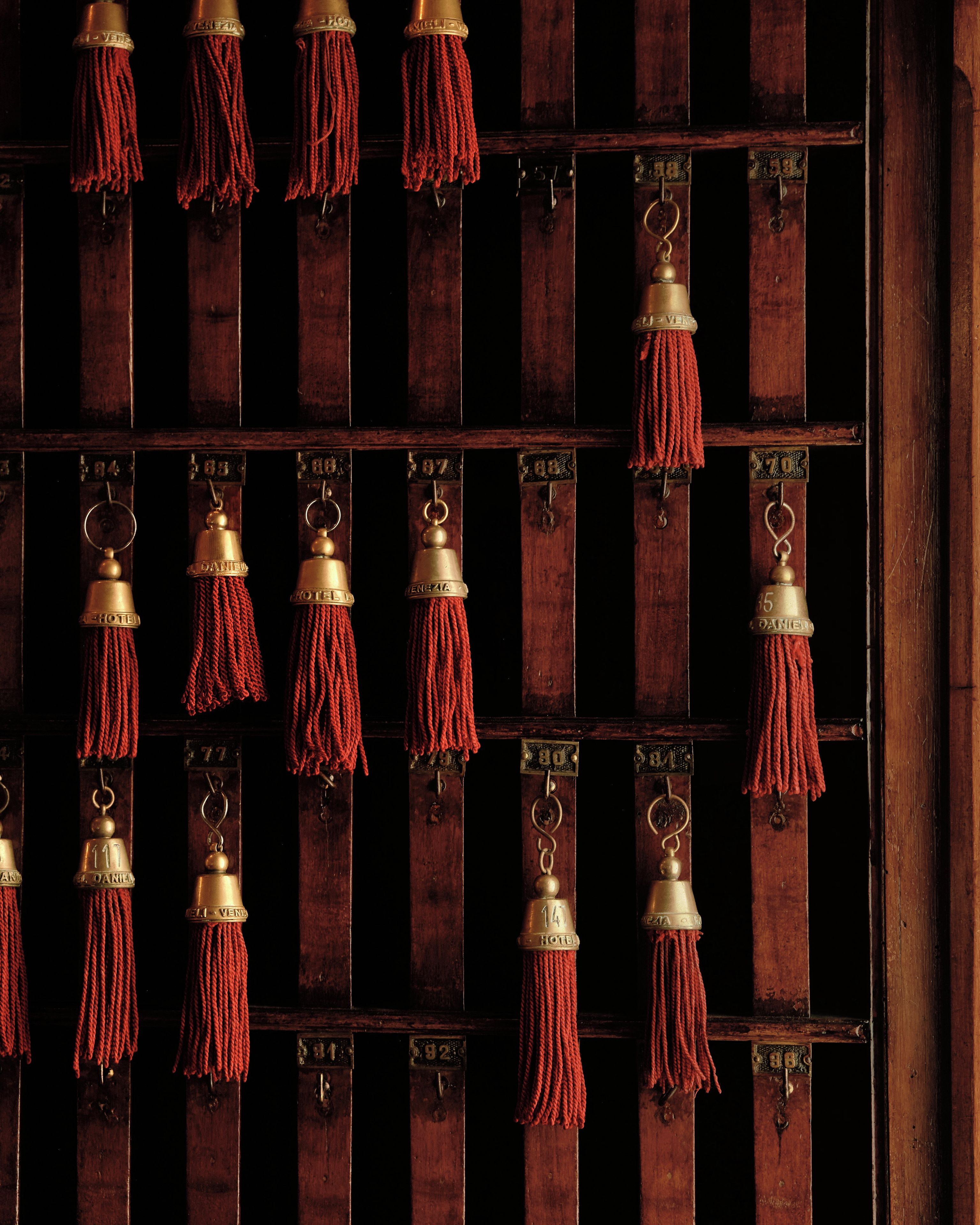 A wall with multiple brass keys hanging on a wooden grid, each with a red tassel and a metal plate with numbers.