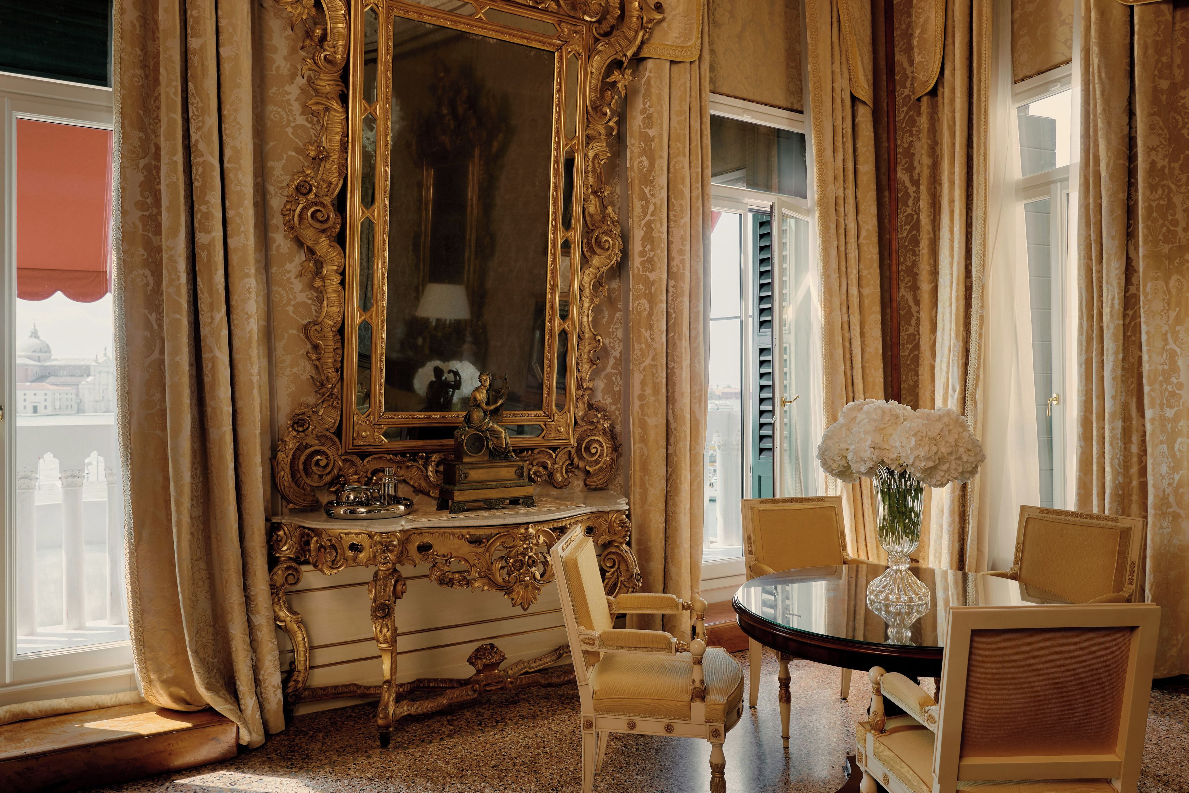 A room with ornate furniture, a large mirror, and a view of a building outside.