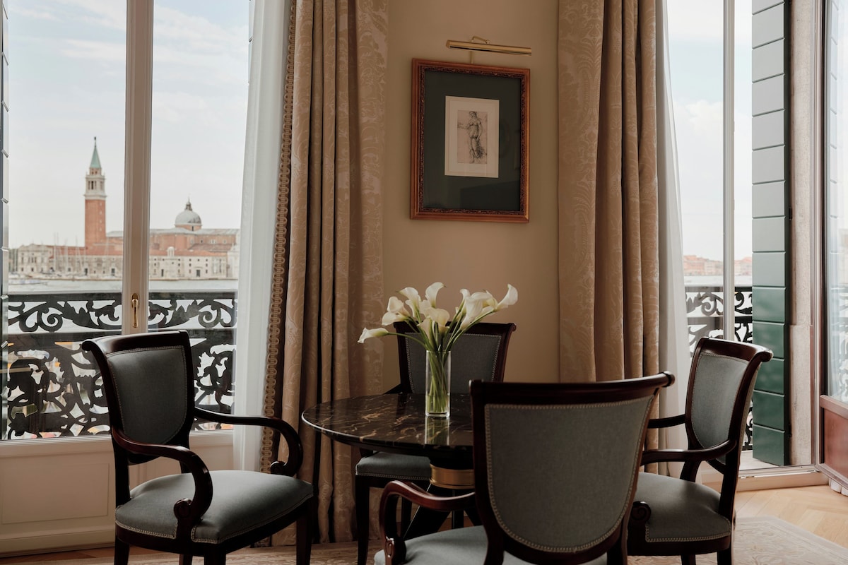Una stanza con un tavolo rotondo, quattro sedie, un vaso con fiori, un quadro appeso al muro e vista sulla città attraverso ampie finestre.