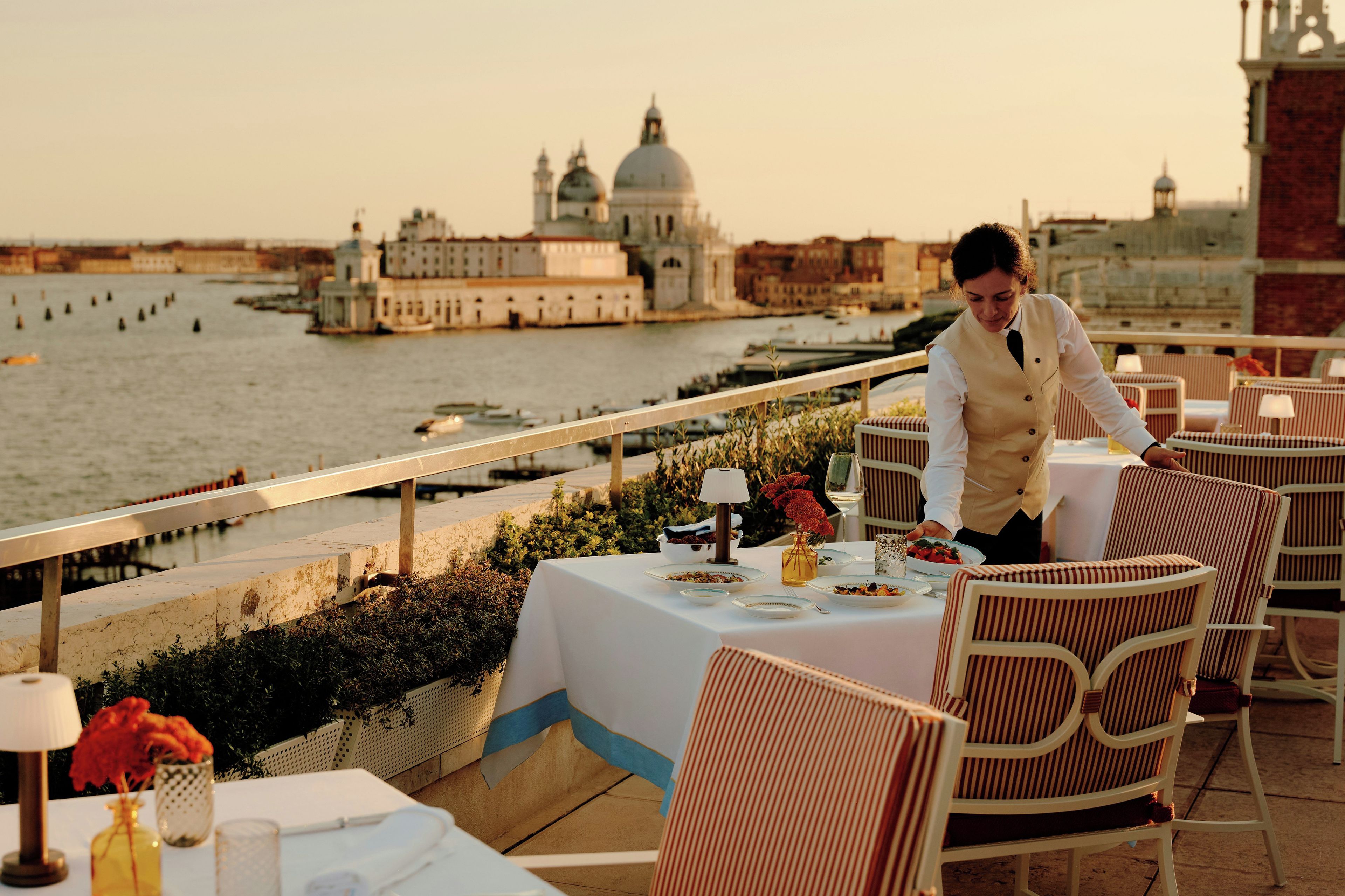 A terrace with a table set for dining, overlooking a body of water with buildings in the background.