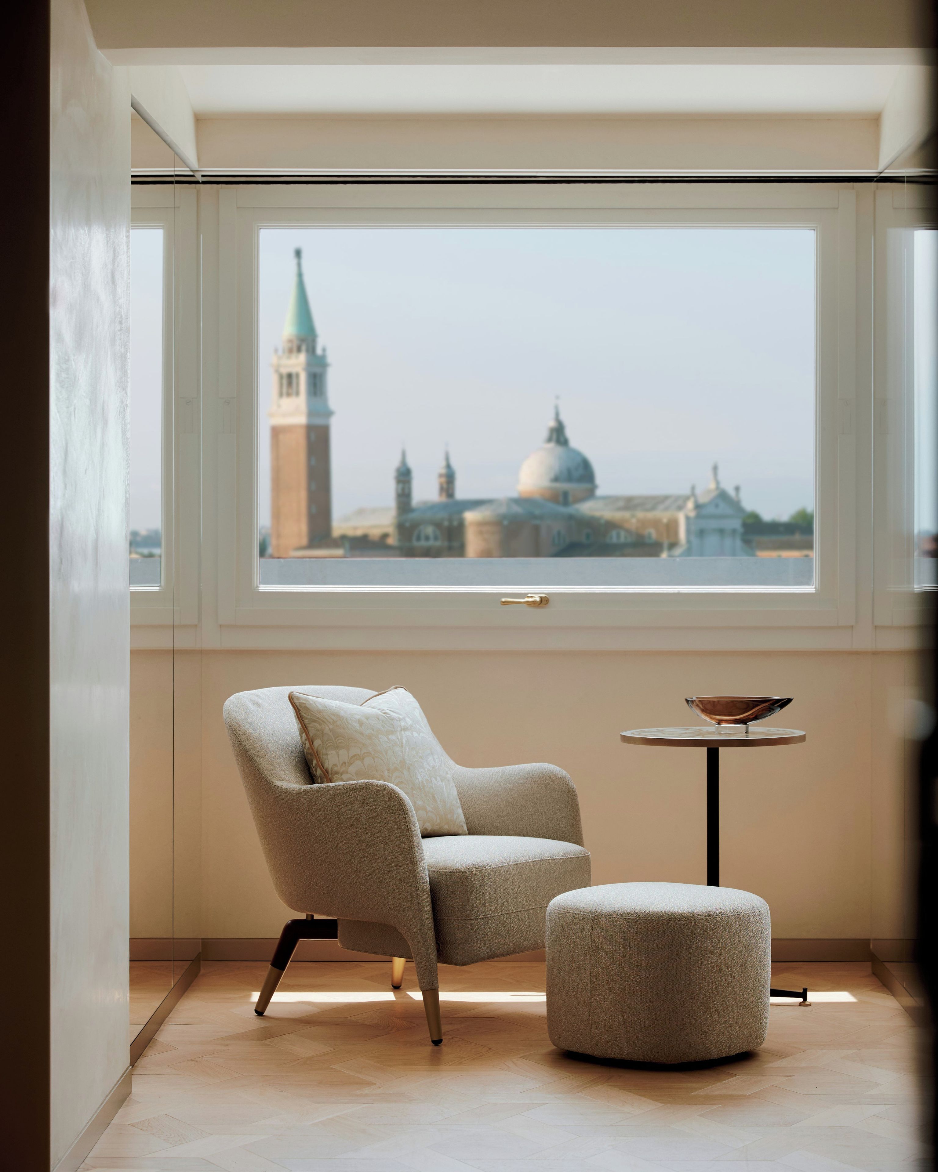 Quarto com vista para paisagem urbana através de uma grande janela, uma cadeira, um banquinho pequeno e uma mesa redonda com um objeto decorativo.