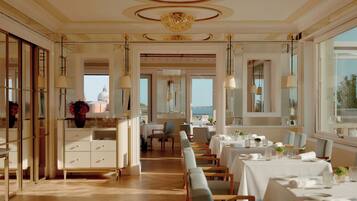 A spacious dining area with high ceilings, elegant chandeliers, and a view of the sea.
