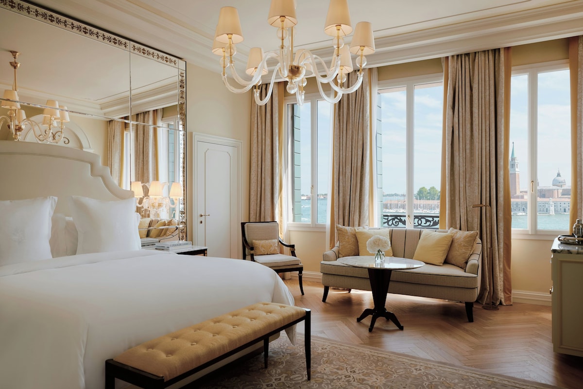 Una lussuosa camera d'hotel con un ampio letto, un'area salotto con divano e poltrone, un tavolino e vista sulla città attraverso grandi finestre.