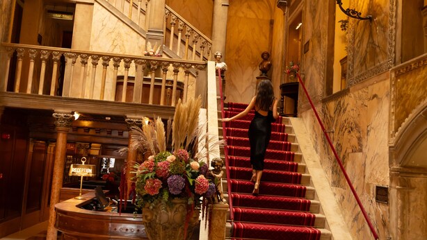 Una majestuosa escalera con alfombra roja, barandillas ornamentadas y un mostrador de recepción con un arreglo floral.