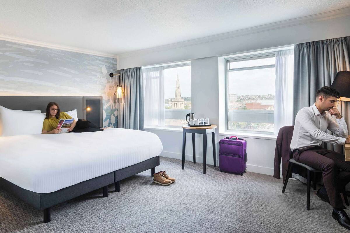 A hotel room with a large bed, a small table with a kettle and cups, a purple suitcase, and a view of a cityscape through the window.
