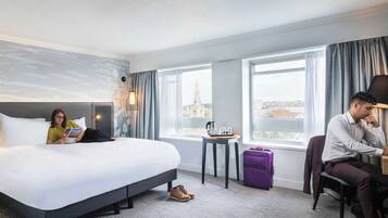 A hotel room with a large bed, a small table with a kettle and cups, a purple suitcase, and a view of a cityscape through the window.