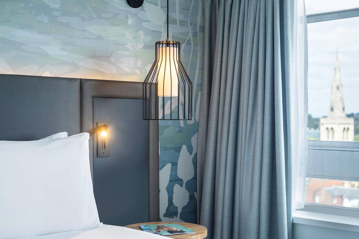 A hotel room with a bed, a nightstand with a lamp, a window with curtains, and a wall with a camouflage pattern.