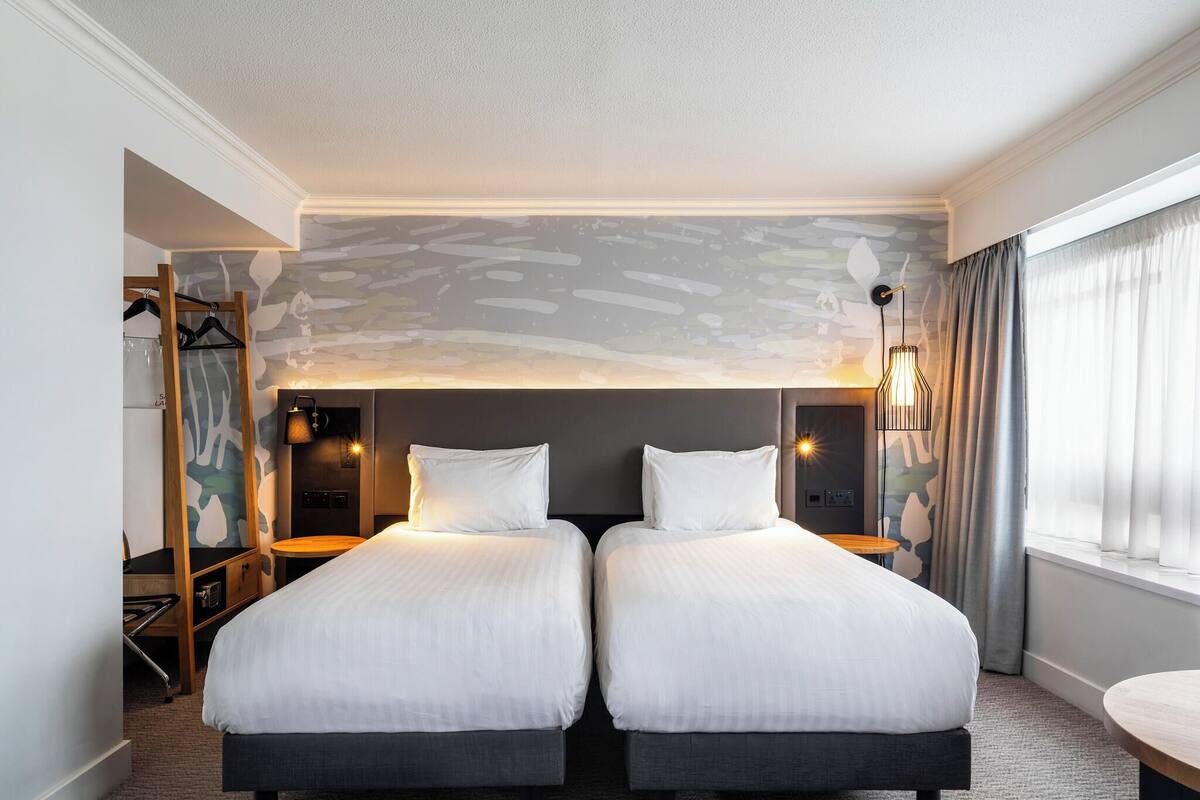 A hotel room with two beds, a small table, a mirror, and a wardrobe.