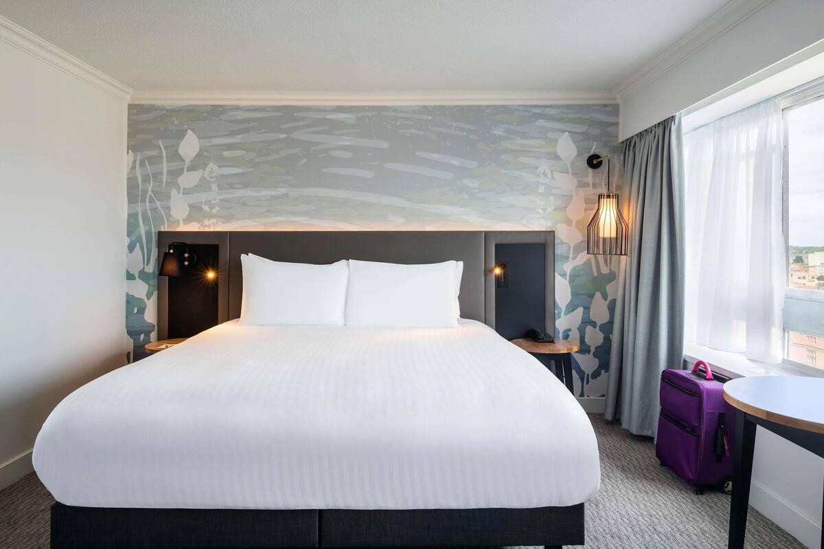 A hotel room with a large bed, a nightstand with a lamp, a suitcase, and a view of the city through the window.