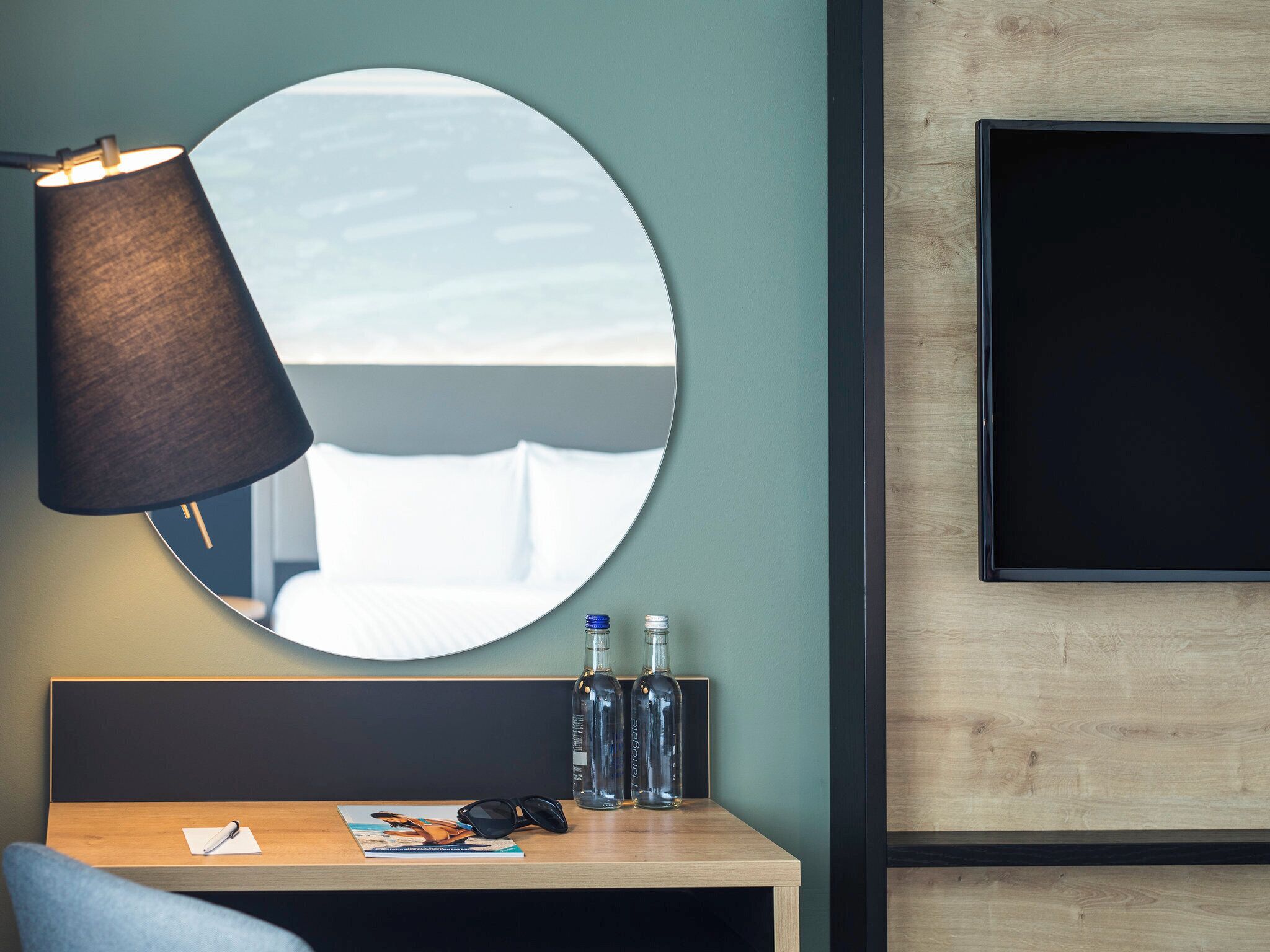 A modern hotel room with a round mirror, a desk with a magazine, a pair of sunglasses, and two bottles of water.
