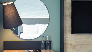 A modern hotel room with a round mirror, a desk with a magazine, a pair of sunglasses, and two bottles of water.