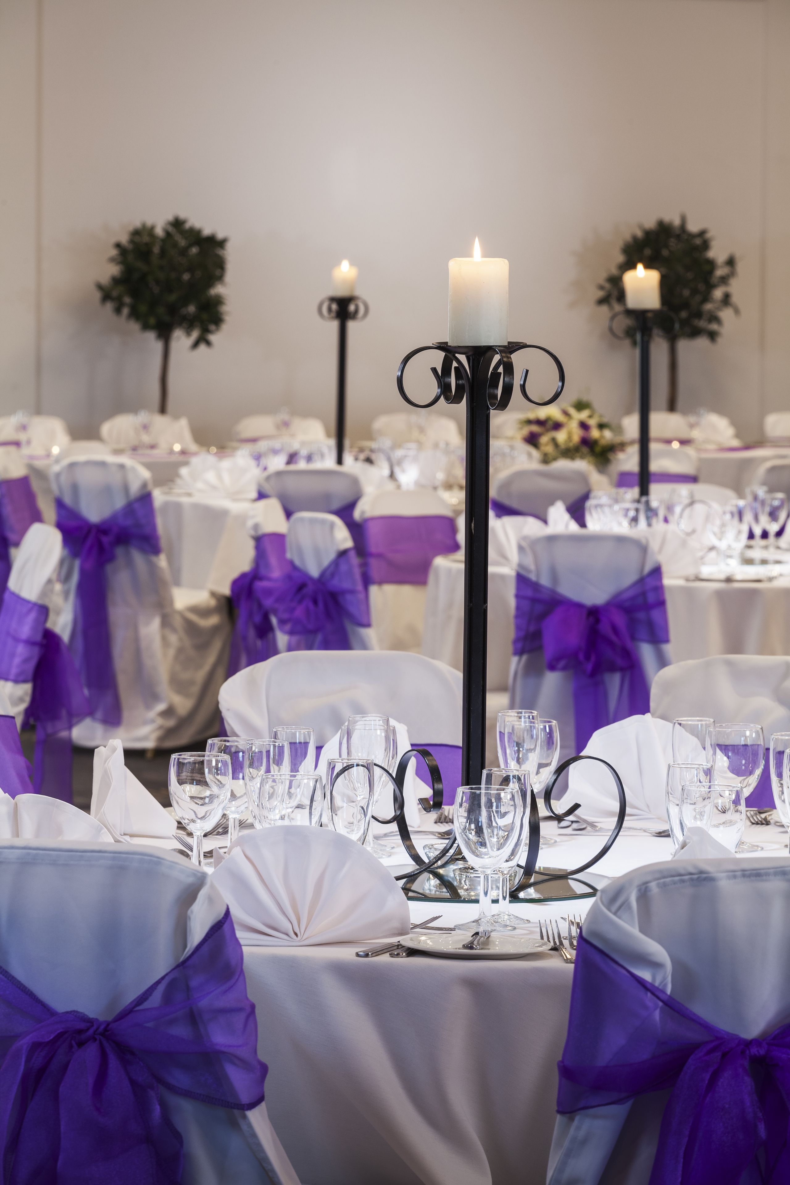 Un salón de banquetes con mesas redondas dispuestas para un evento formal, decorado con manteles blancos y cintas moradas.