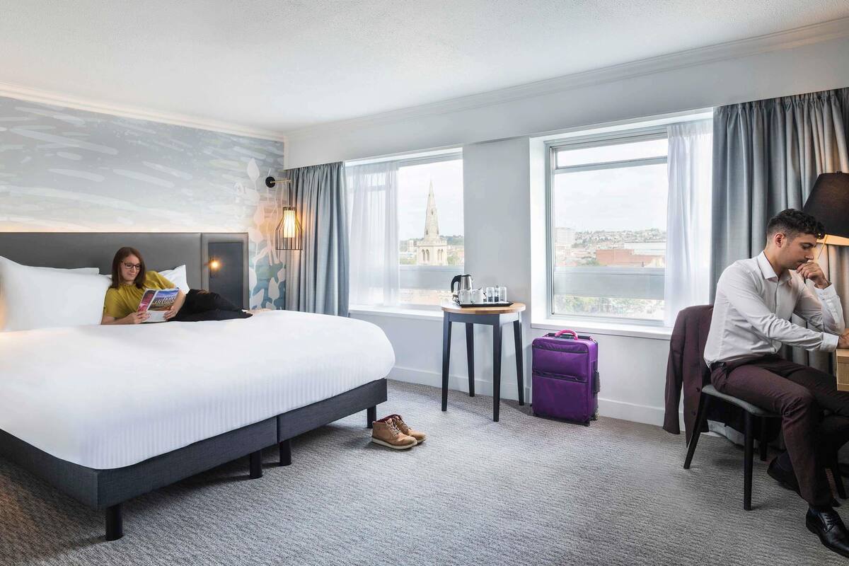 A hotel room with a bed, a small table with a kettle, a suitcase, and a view of a cityscape.