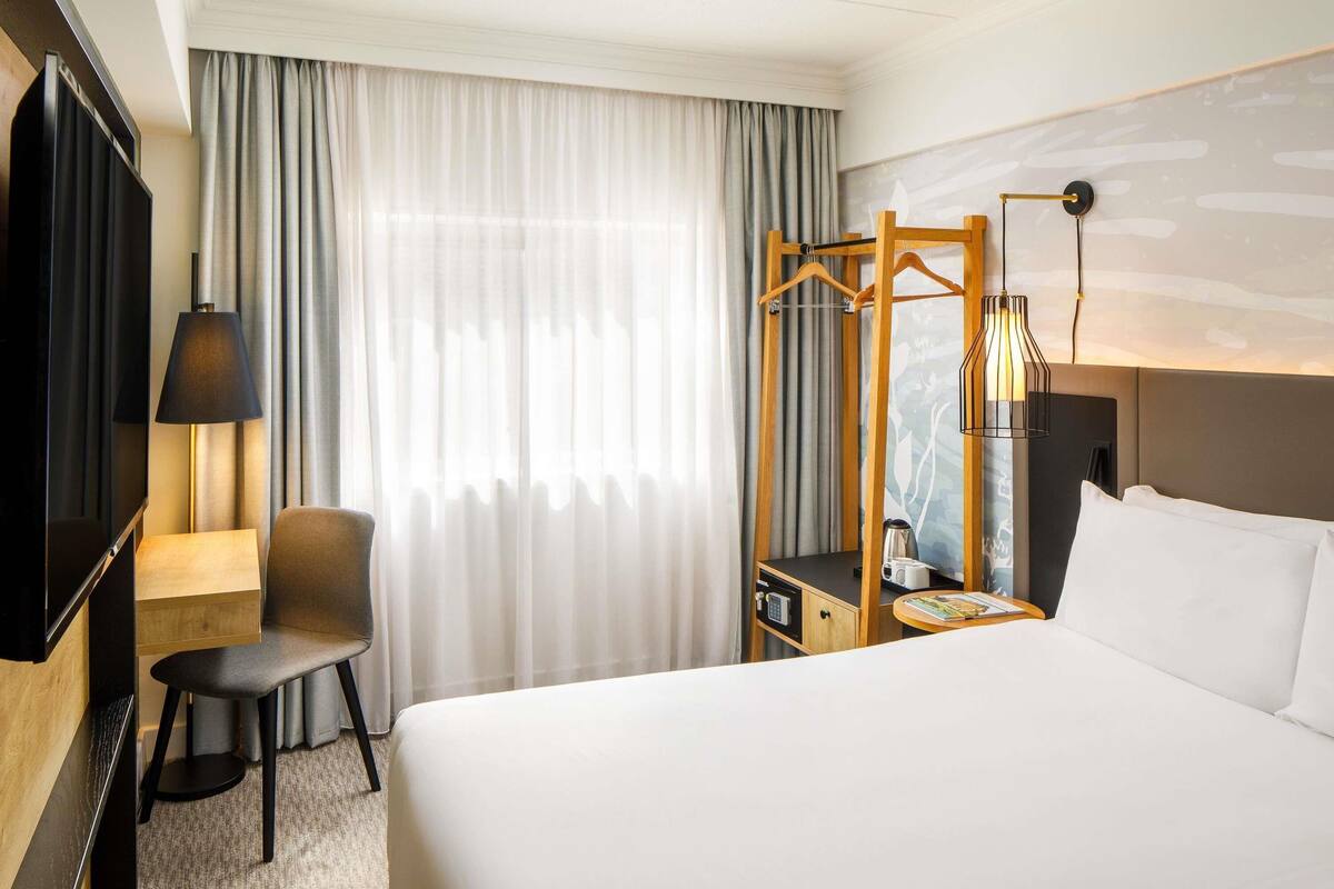 A hotel room with a bed, a desk with a chair, a TV, a lamp, a coat rack, and a bedside table with a kettle.