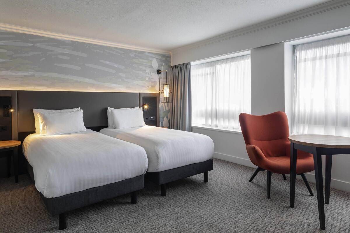 A hotel room with two beds, a chair, a table, and a large window with curtains.