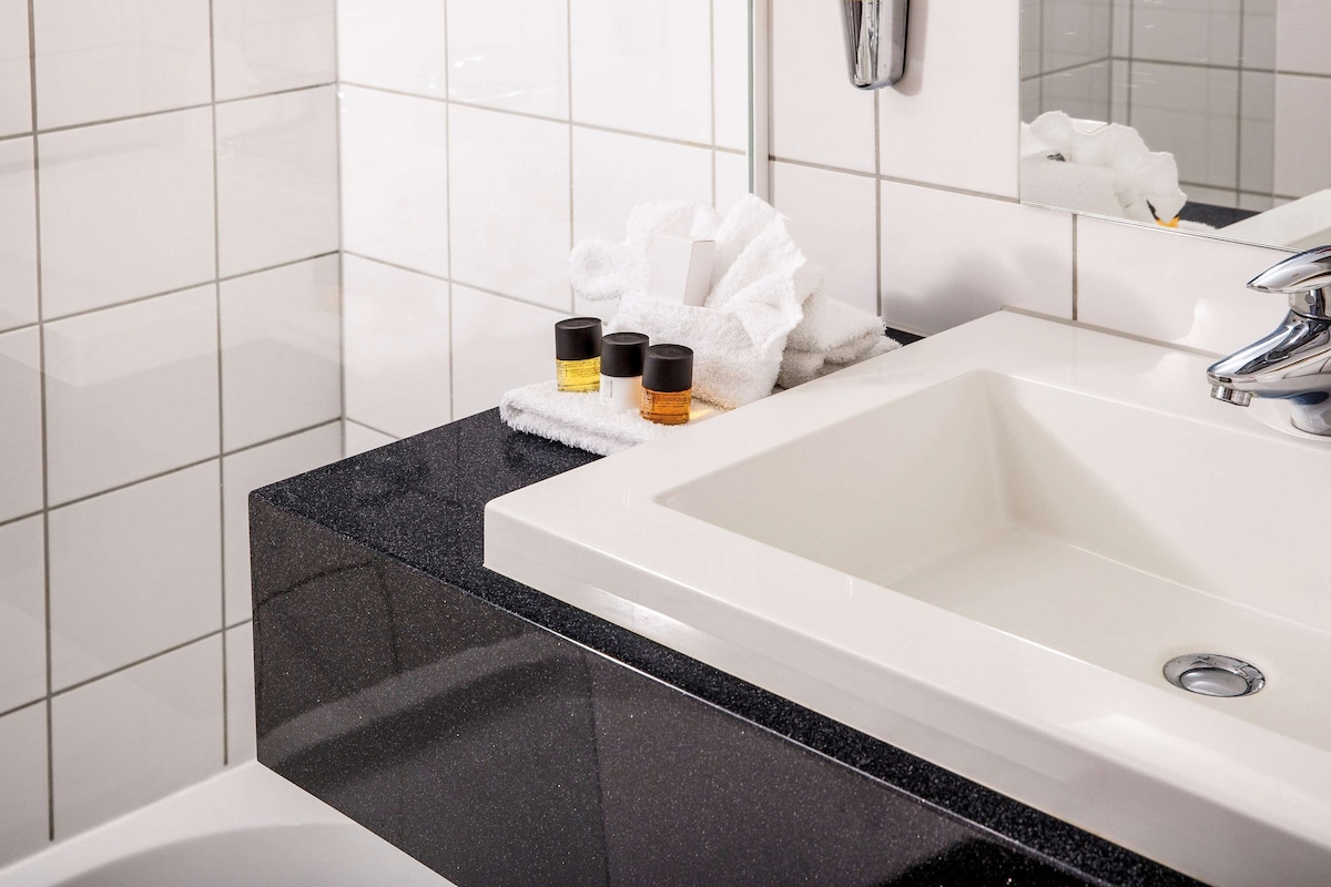 Une salle de bain avec un lavabo blanc, un plan de toilette noir et des murs recouverts de carreaux blancs. On trouve des articles de toilette sur le plan de toilette et une serviette accrochée au mur.