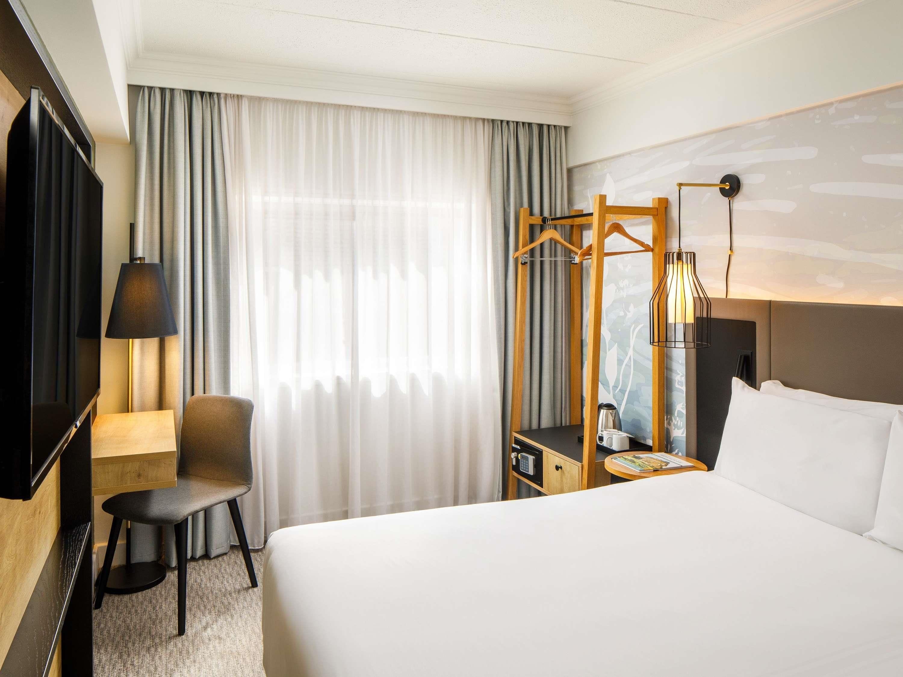 A hotel room with a bed, a desk, a chair, a TV, a lamp, a coat rack, and a window with curtains.