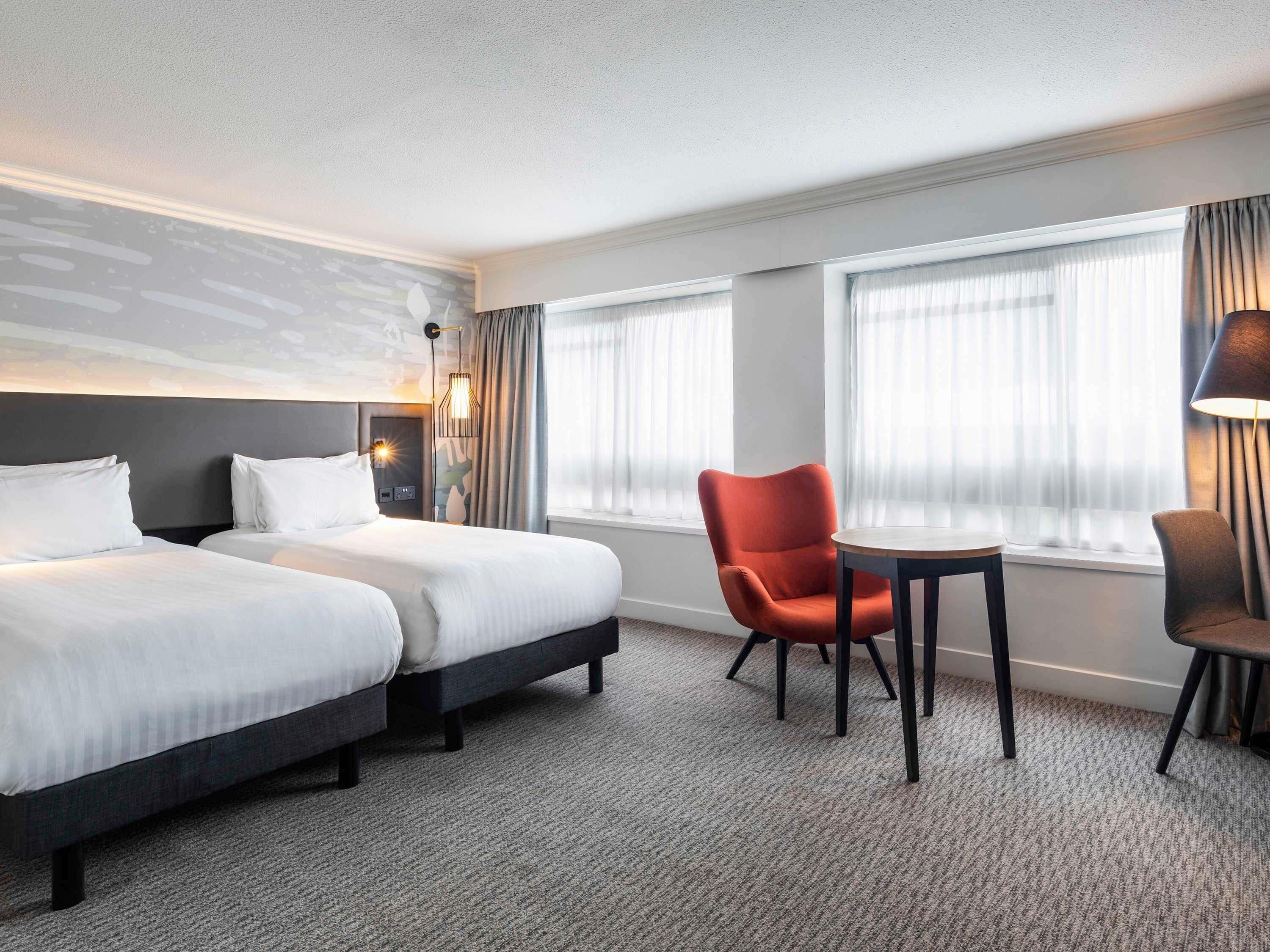 A hotel room with two beds, a small table, a chair, and a lamp.