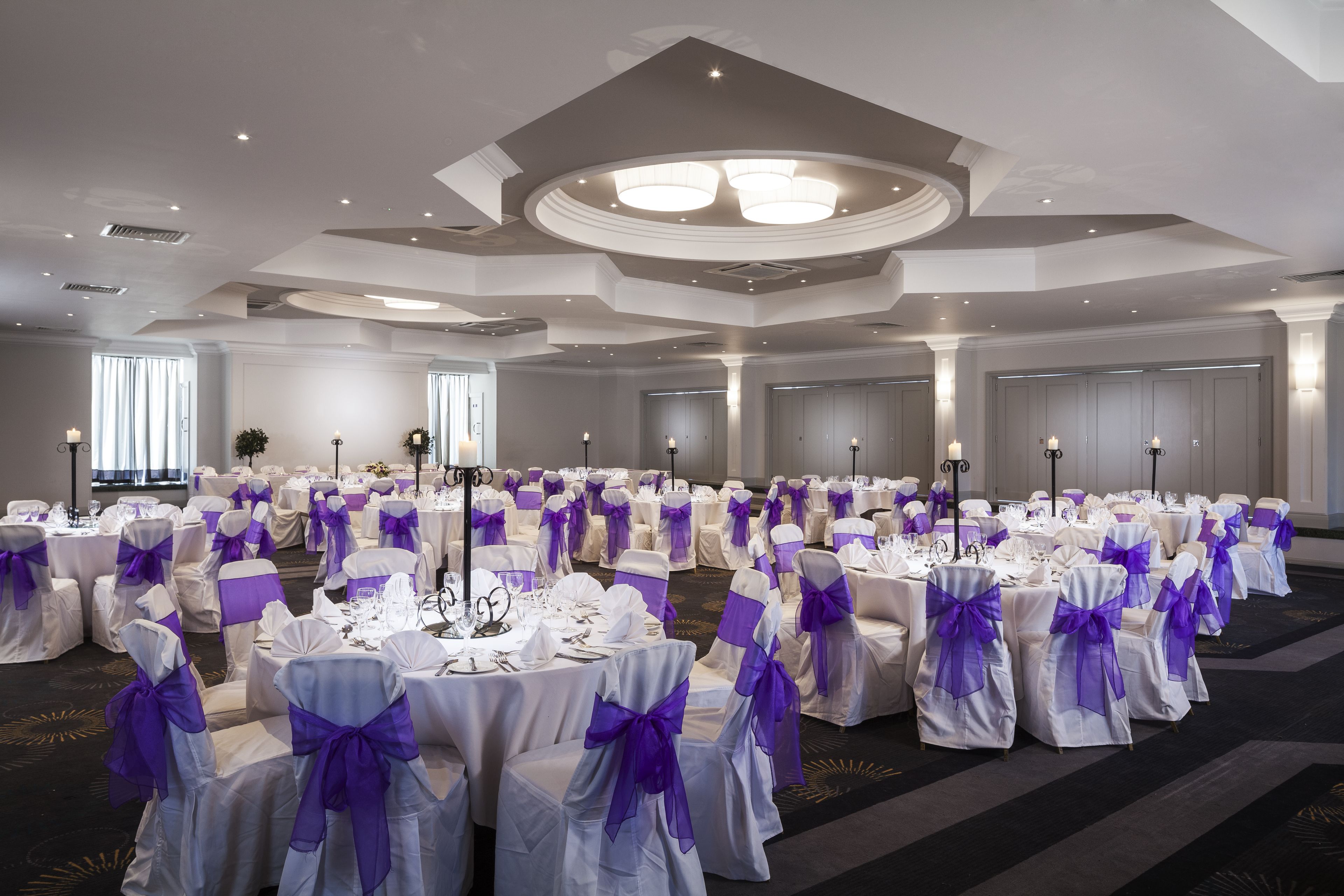 Un gran salón de banquetes con mesas redondas y sillas, cada una decorada con manteles blancos y fajas moradas.